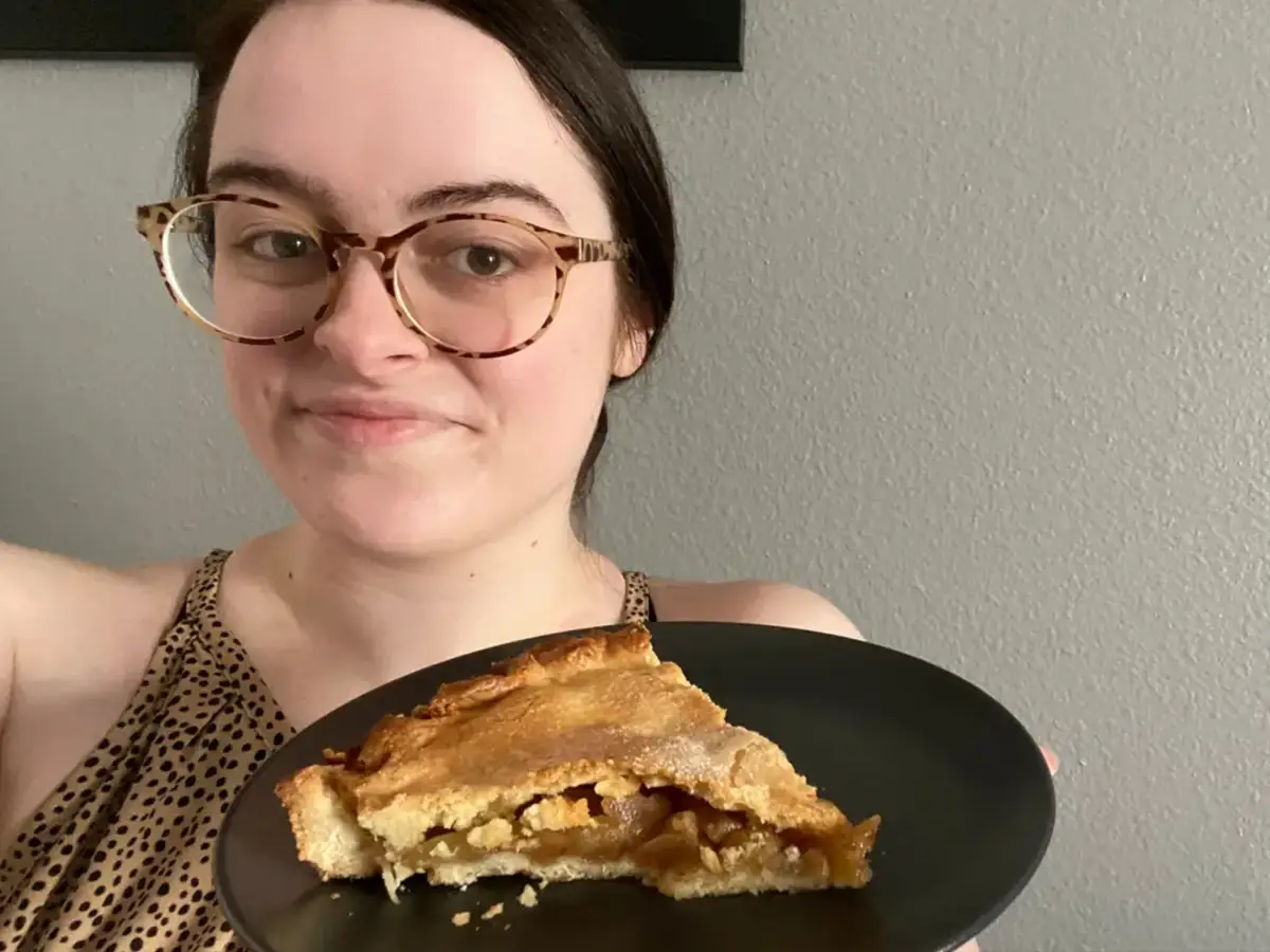 Selfie of the writer Paige Bennett against a white wall holding a gray plate with a slice of apple pie on it
