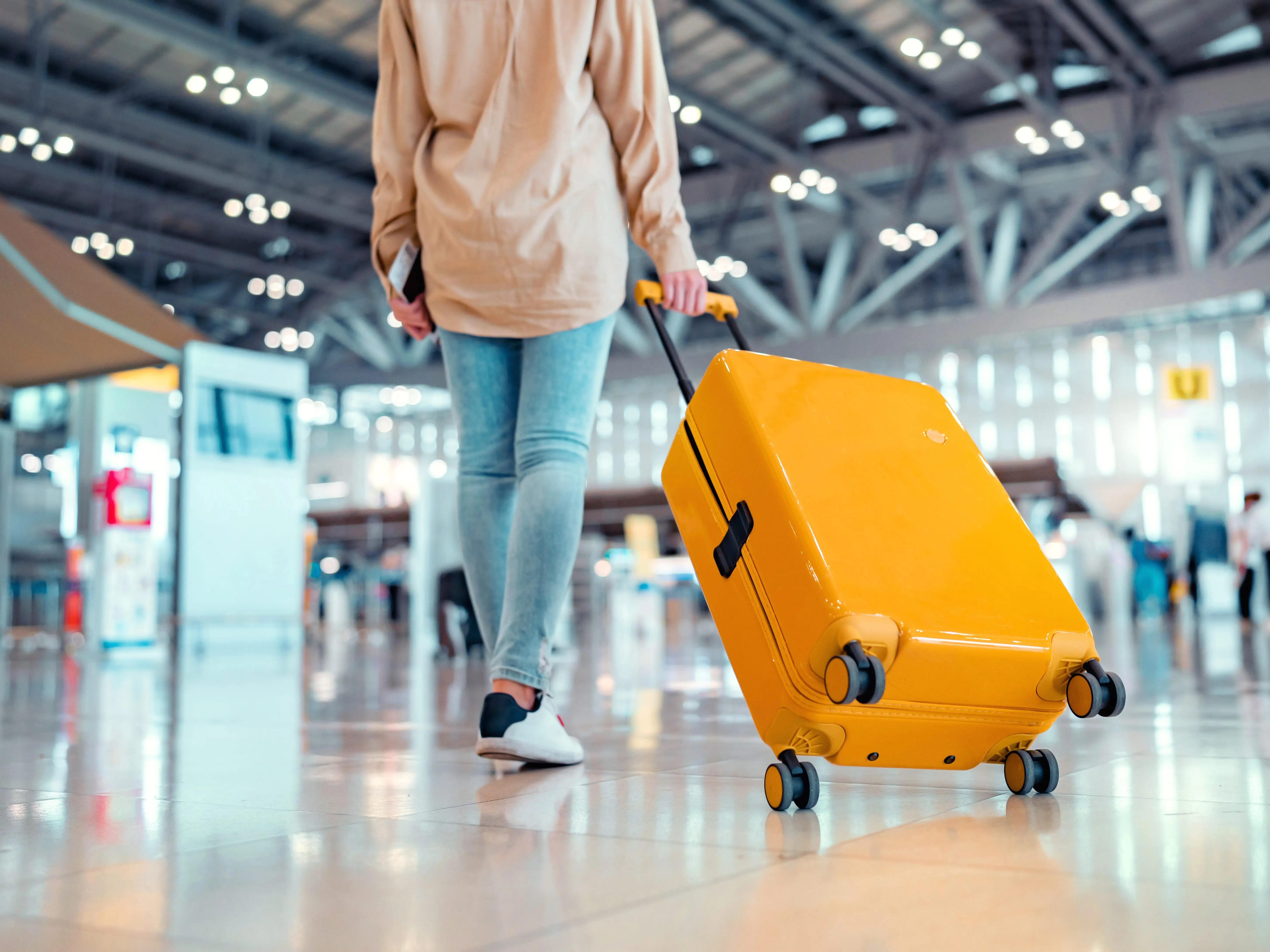 A woman walking through the airport carrying a passport, ticket, and bright yellow suitcase.