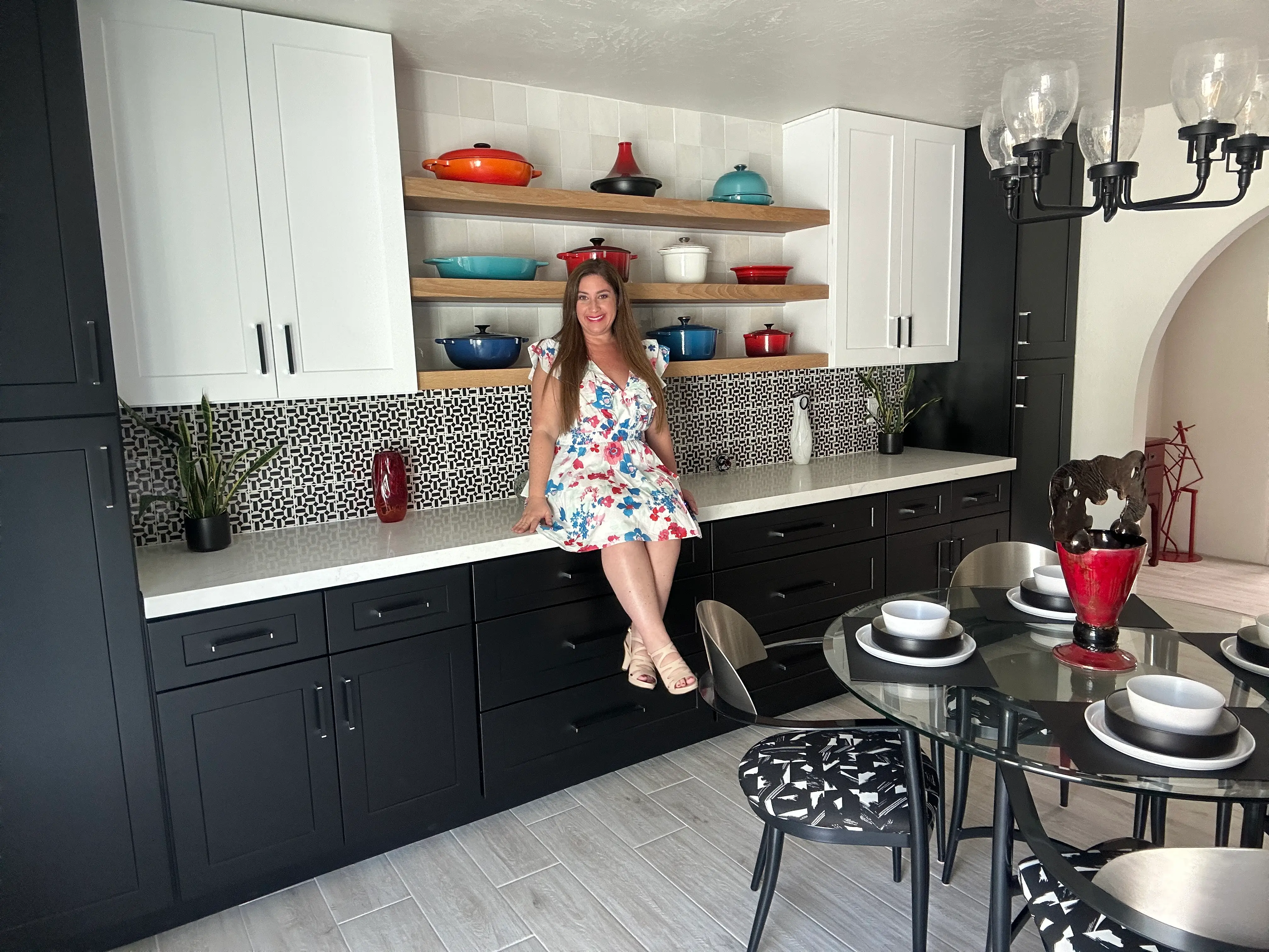 Author Jill Schildhouse sitting on counter with white marble tops, black cabinets, and black harware with shelf of colorful pots behind her on open shelving