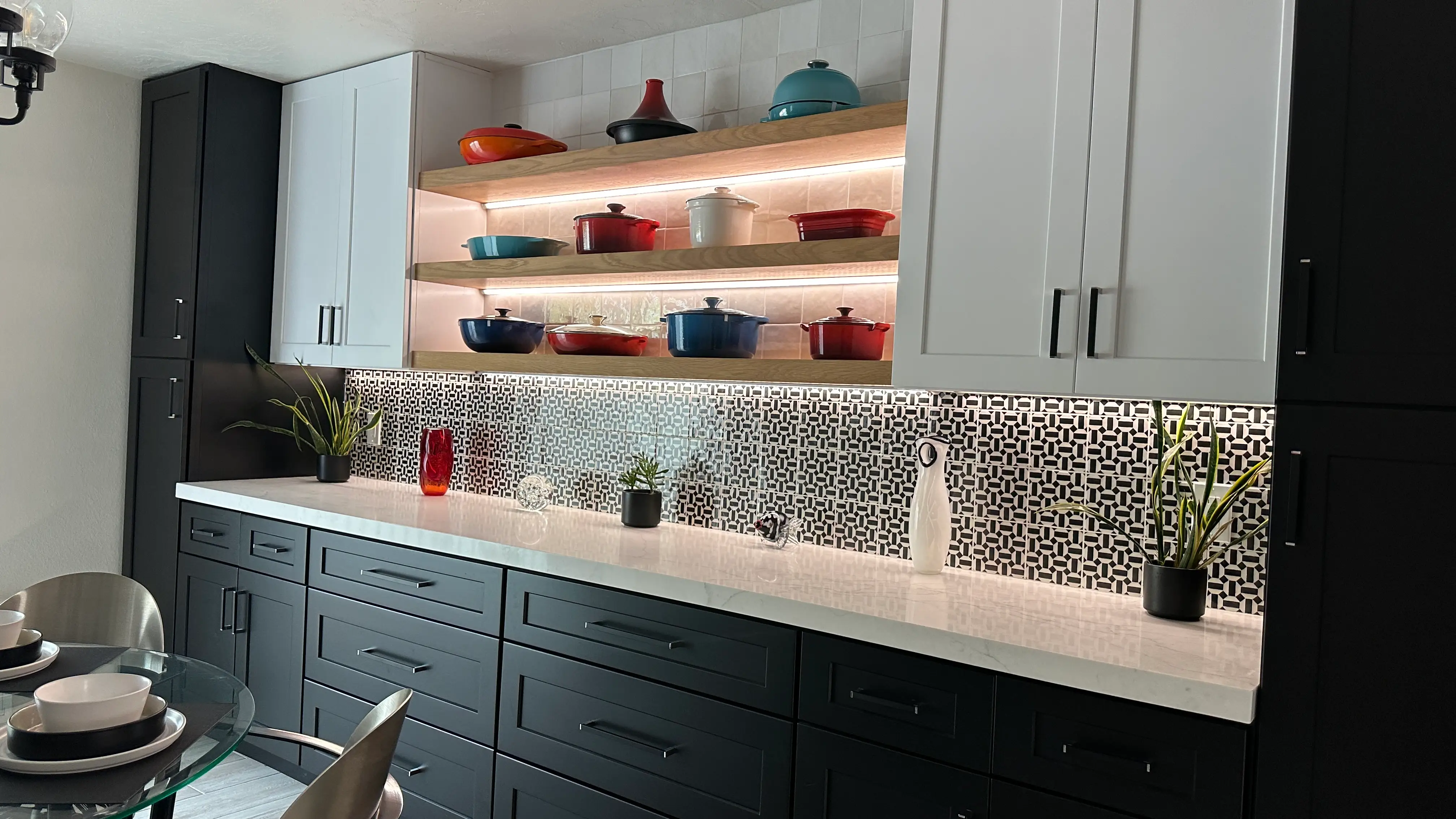 Back-lit open shelves with colorful cookingware above black cabinets with black hardware and white countertops with a patterned geometric black and white backsplash and white cabinets with black hardware above it