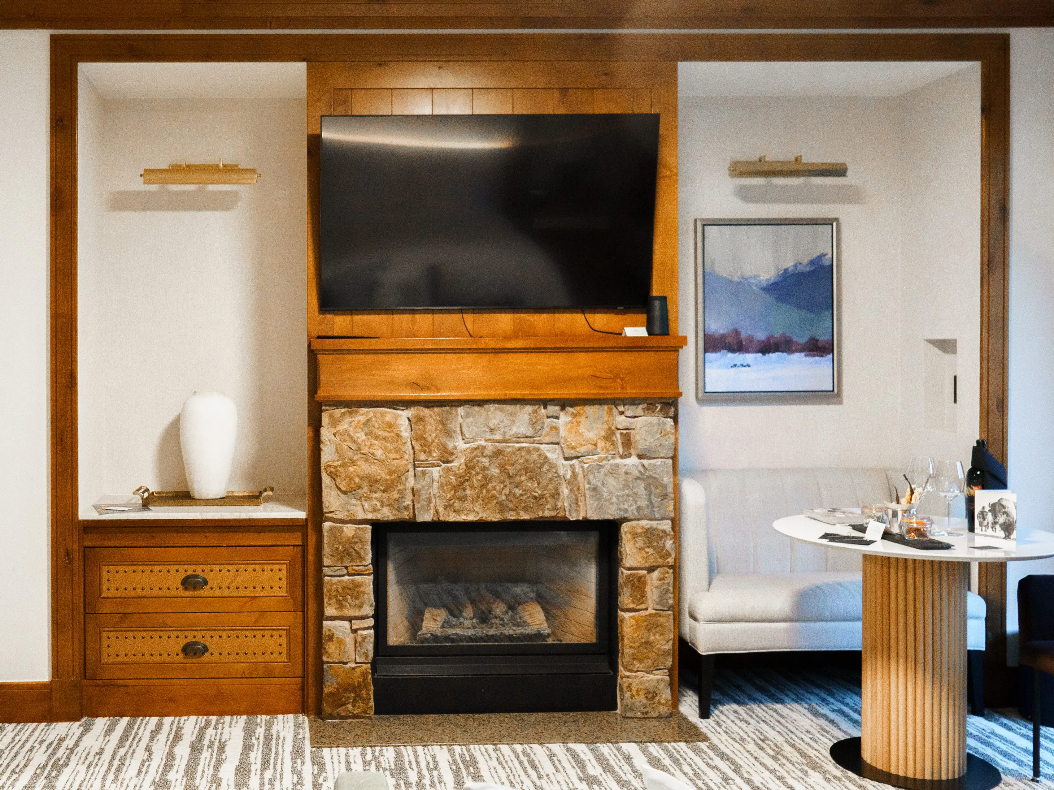 Inside a hotel room with white walls and wood trimmings, a table and chairs on the right, and a TV mounted above a stone fireplace in the center