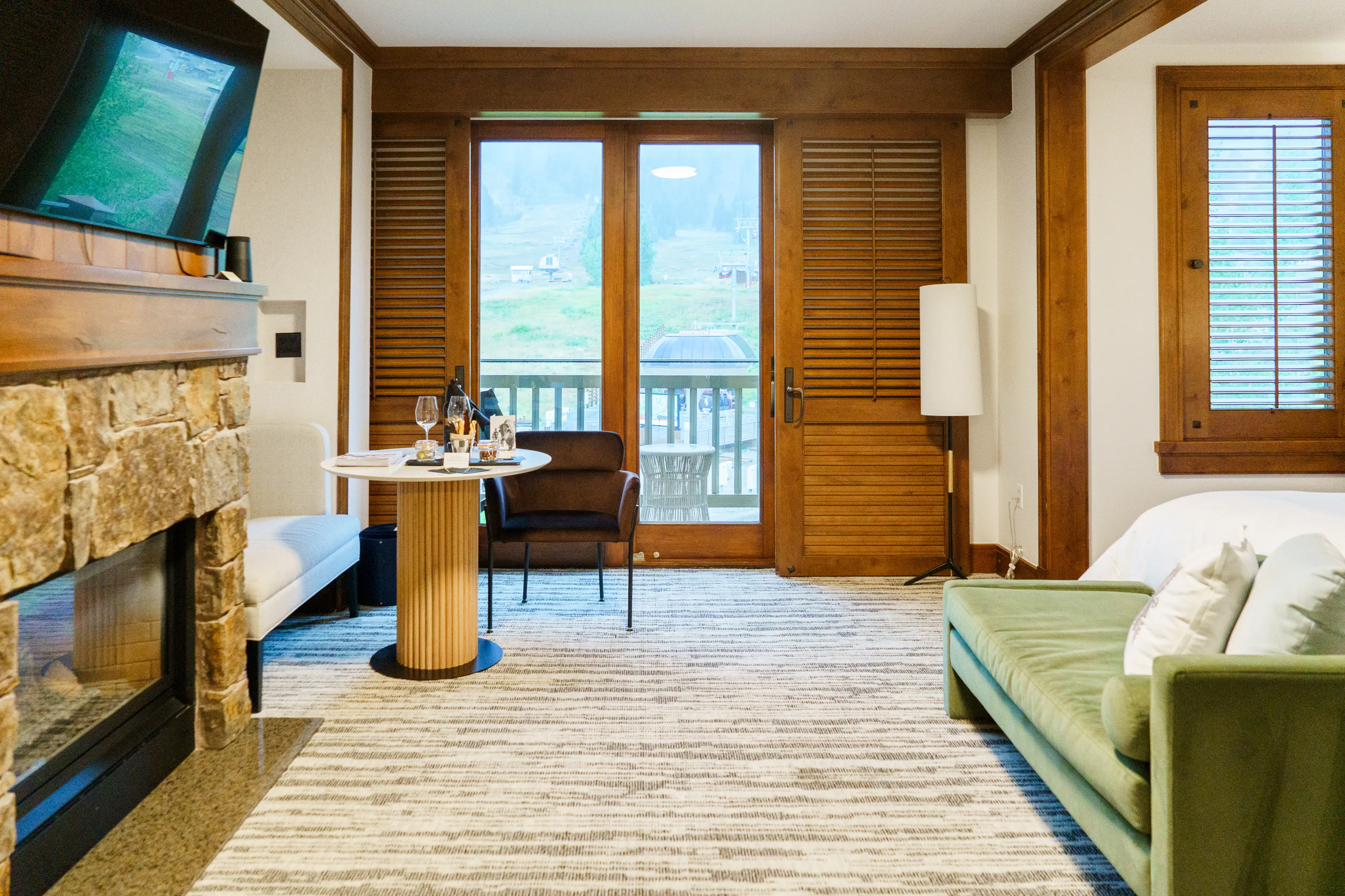 Inside a hotel room with a TV above a fireplace on the left, a green couch on the right, and wood shutters leading to a balcony in the back