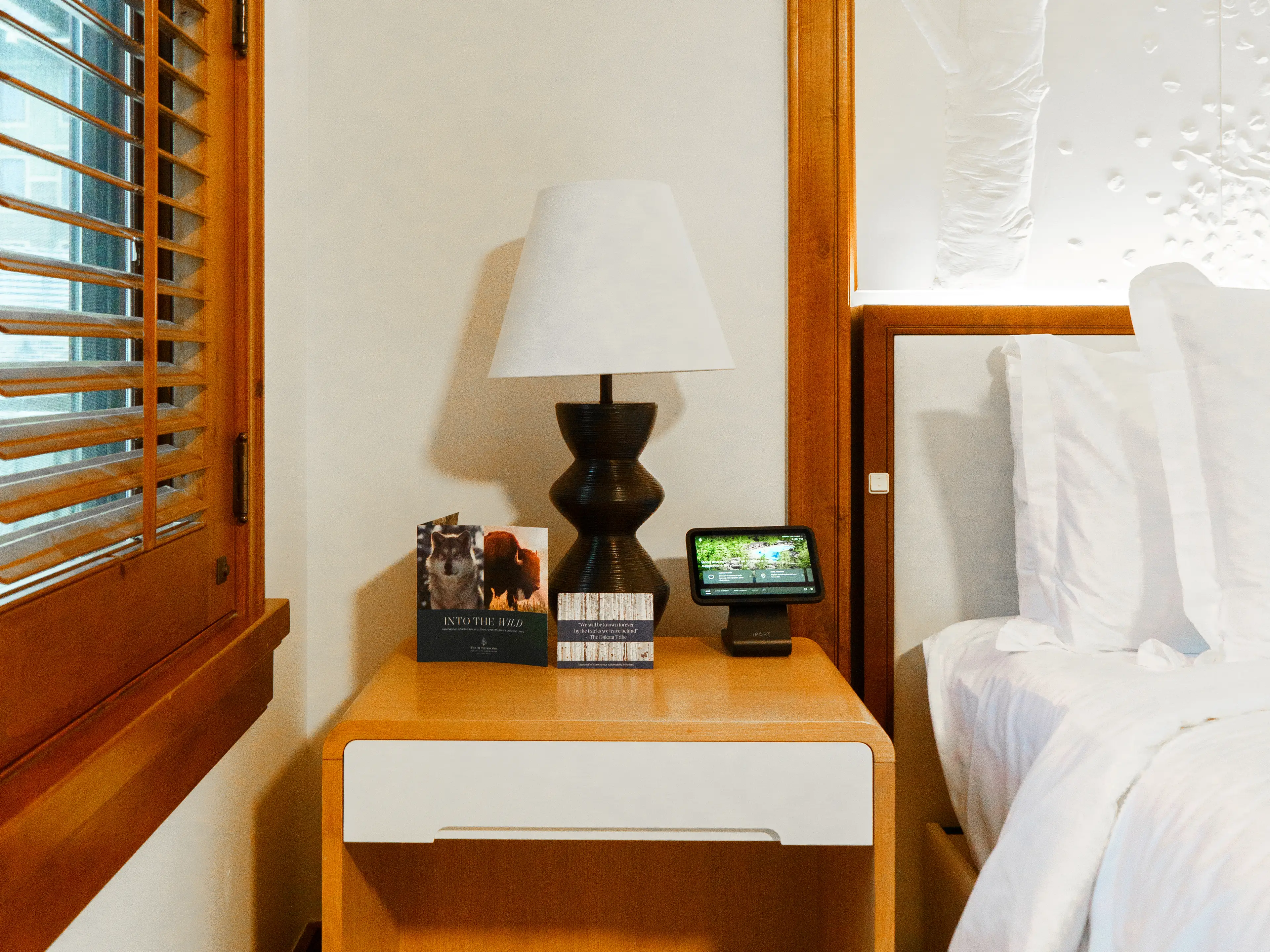 A wooden nightstand between a bed and shutterd windows holds two brochures and a tablet on a stand in front of a modern lamp