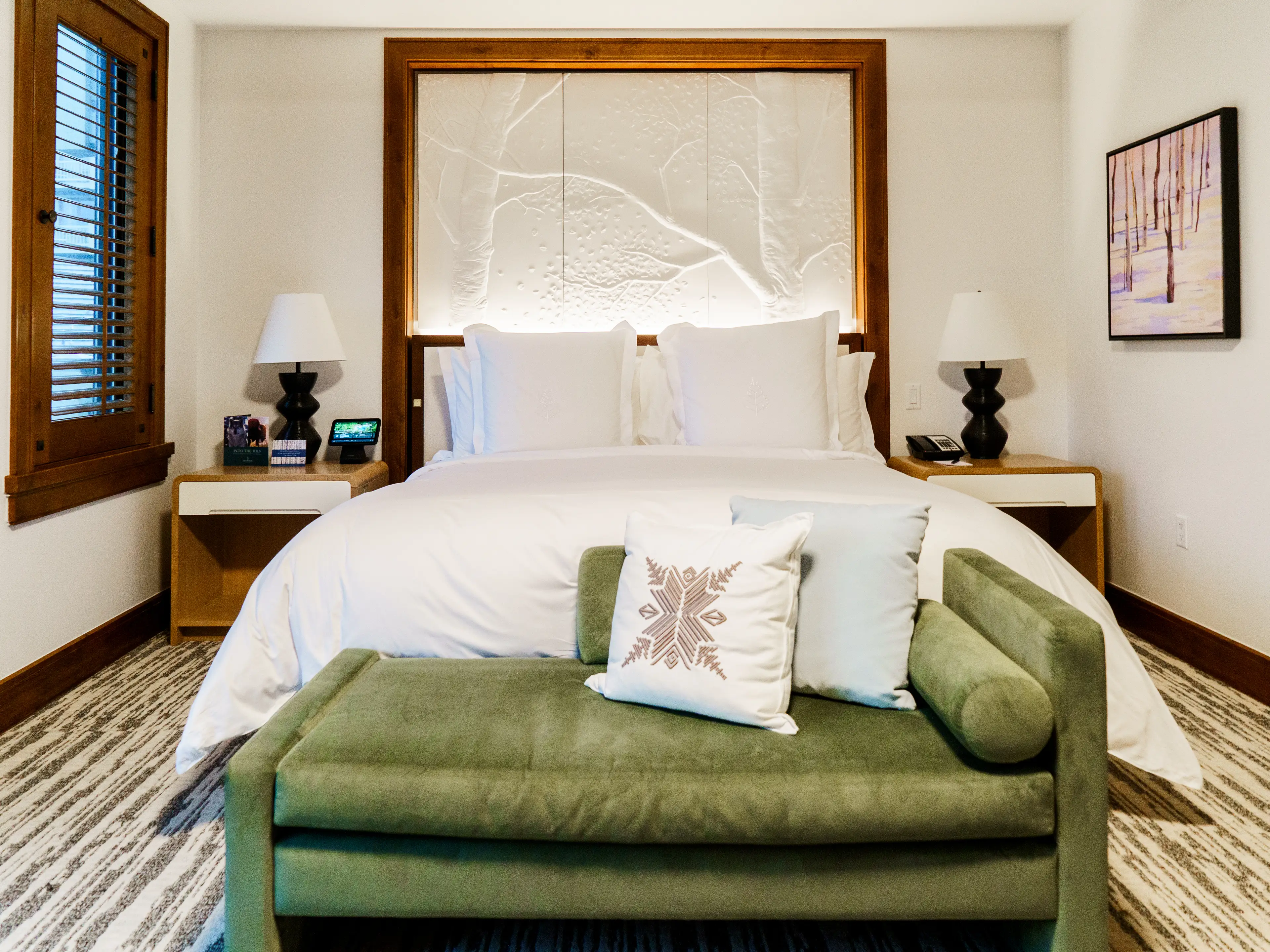 Inside a hotel room with a green couch in front of a white bed book-ended by nightstands with lamps on them, and a large white headboard with tree carvings on it