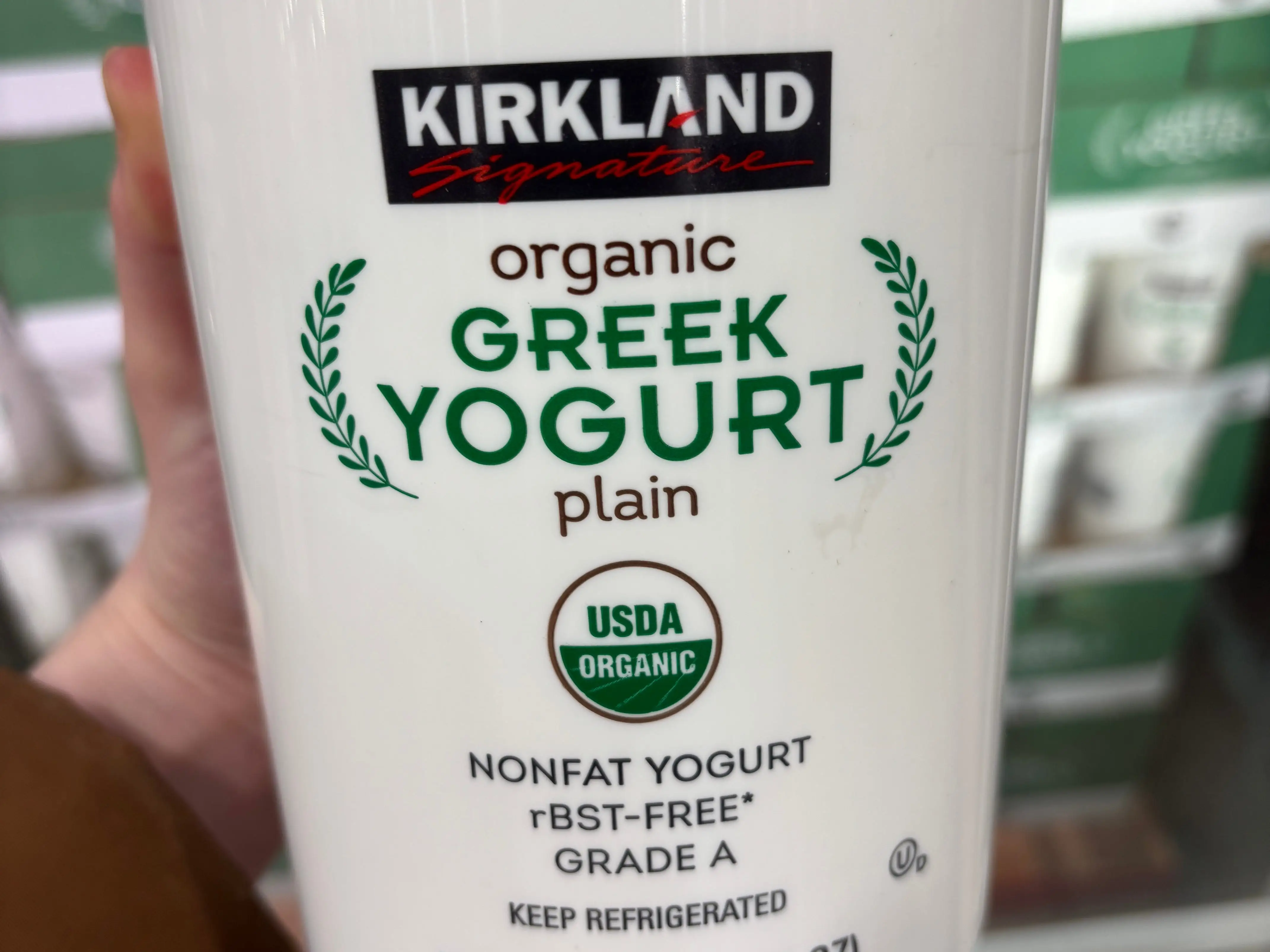 A hand holding a container of Kirkland Signature organic plain Greek yogurt.