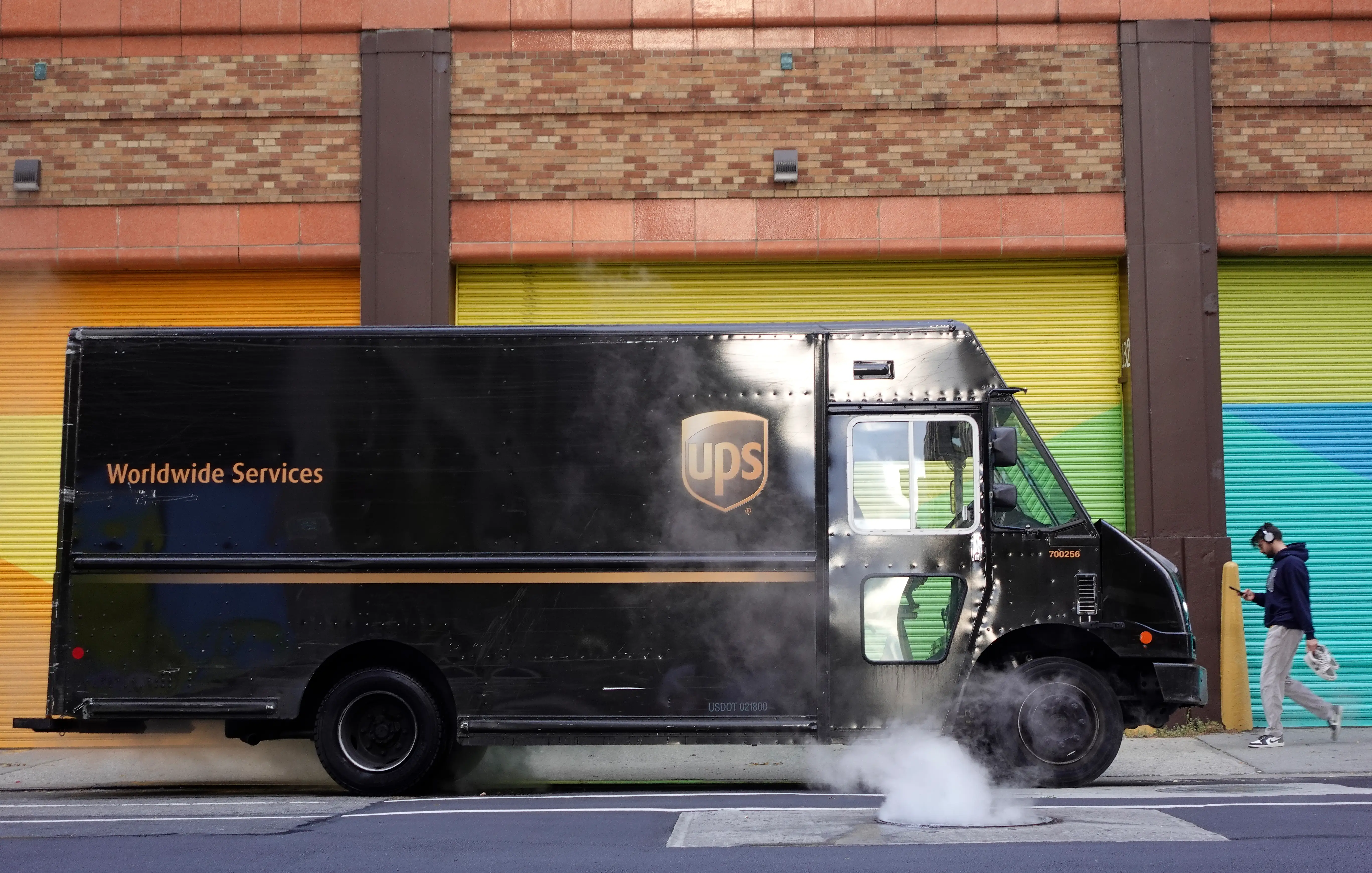 A dark brown UPS truck sits parked on a street as steam rises from a manhole next to it and a pedestrian walks by on the sidewalk wearing headphones and holding a smartphone.