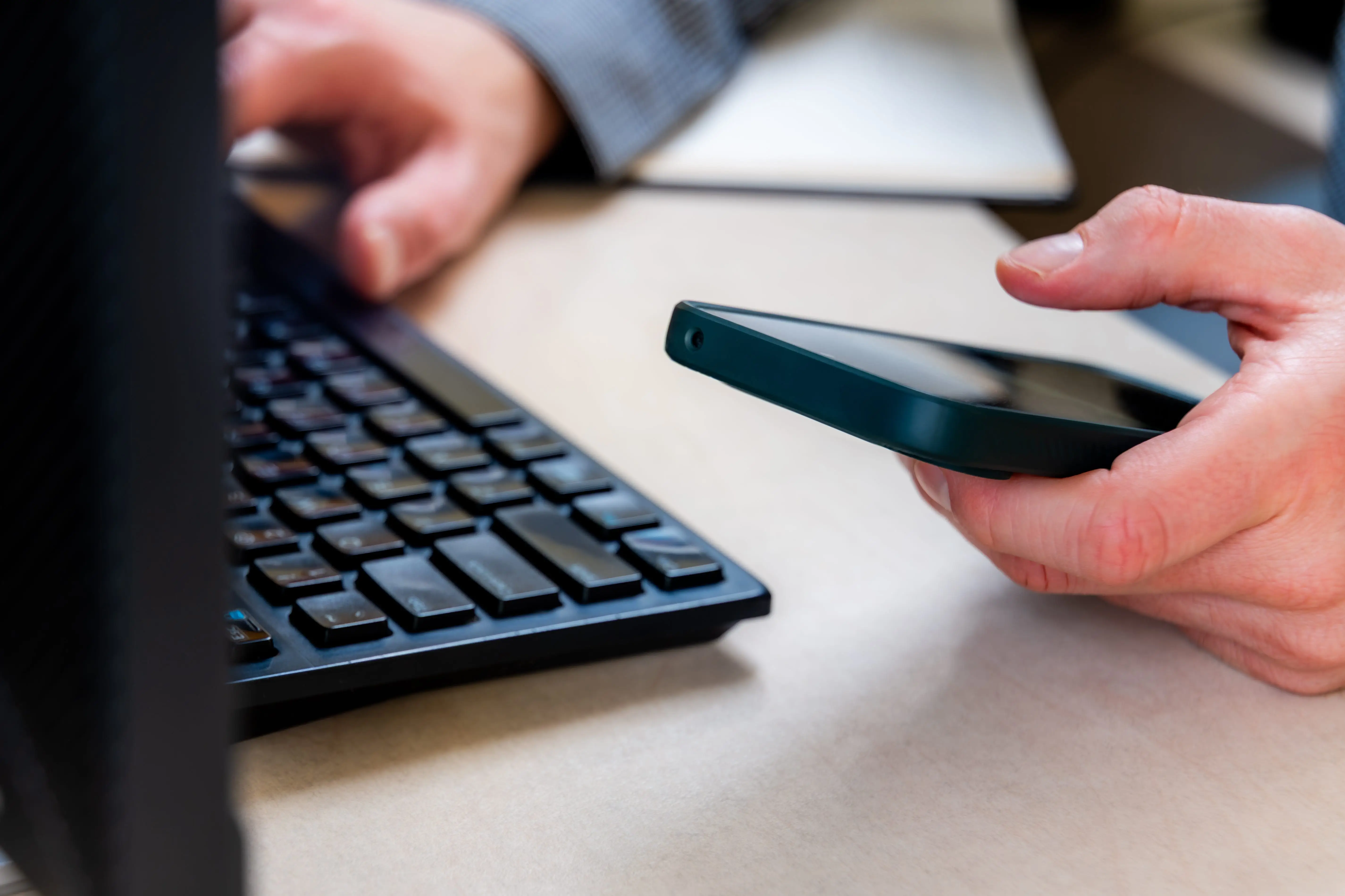 Hand Holds Smartphone Near Computer Keyboard At Desk, Showing Multitasking Communication, Notifications, And Mobile Work Updates For Business Productivity In A Modern Office Workflow.