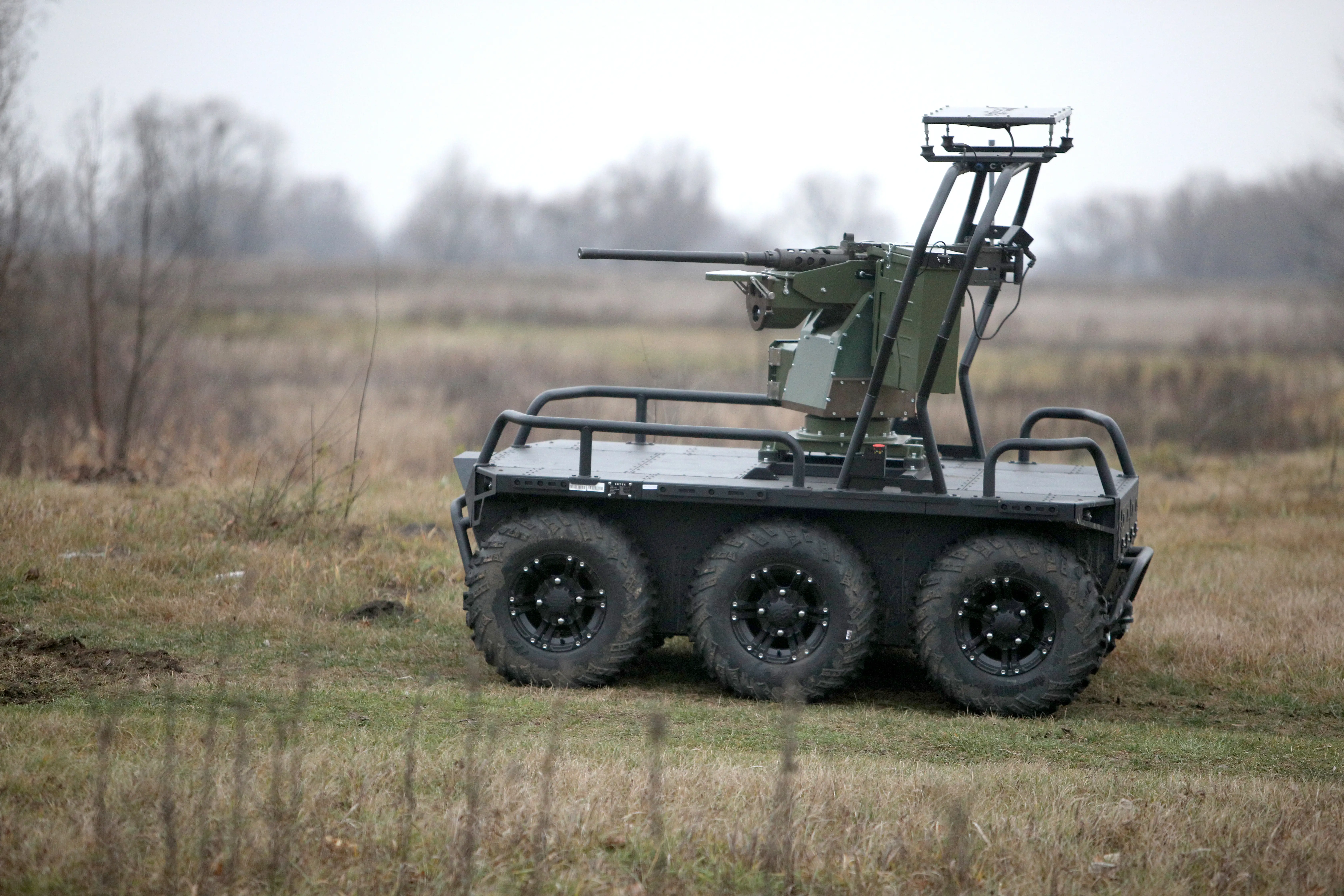 A green robot on six wheels with a machine gun on top and shubbery around it