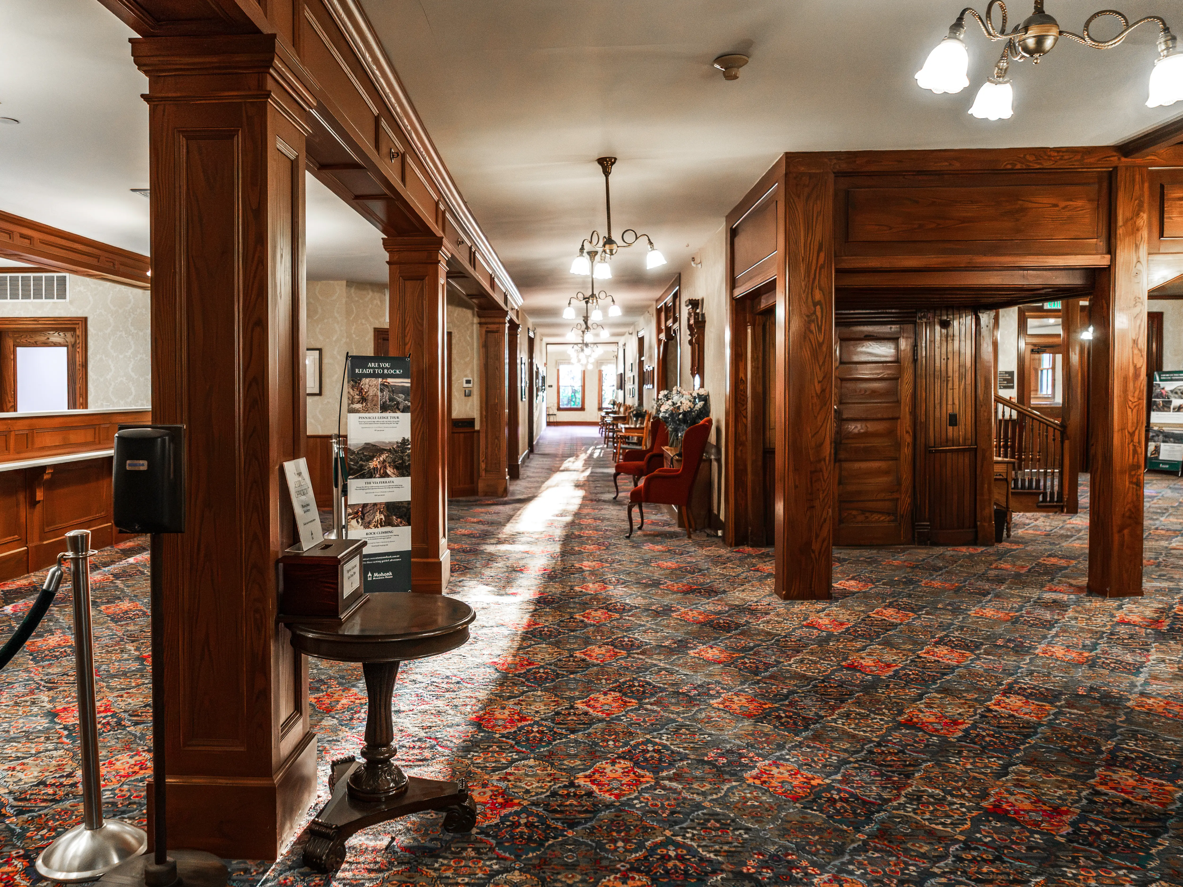 Inside the lobby at Mohonk Mountain House is a wood-paneled corridor with patterned carpet, chandeliers, seating, and display signs in an ornate interior.
