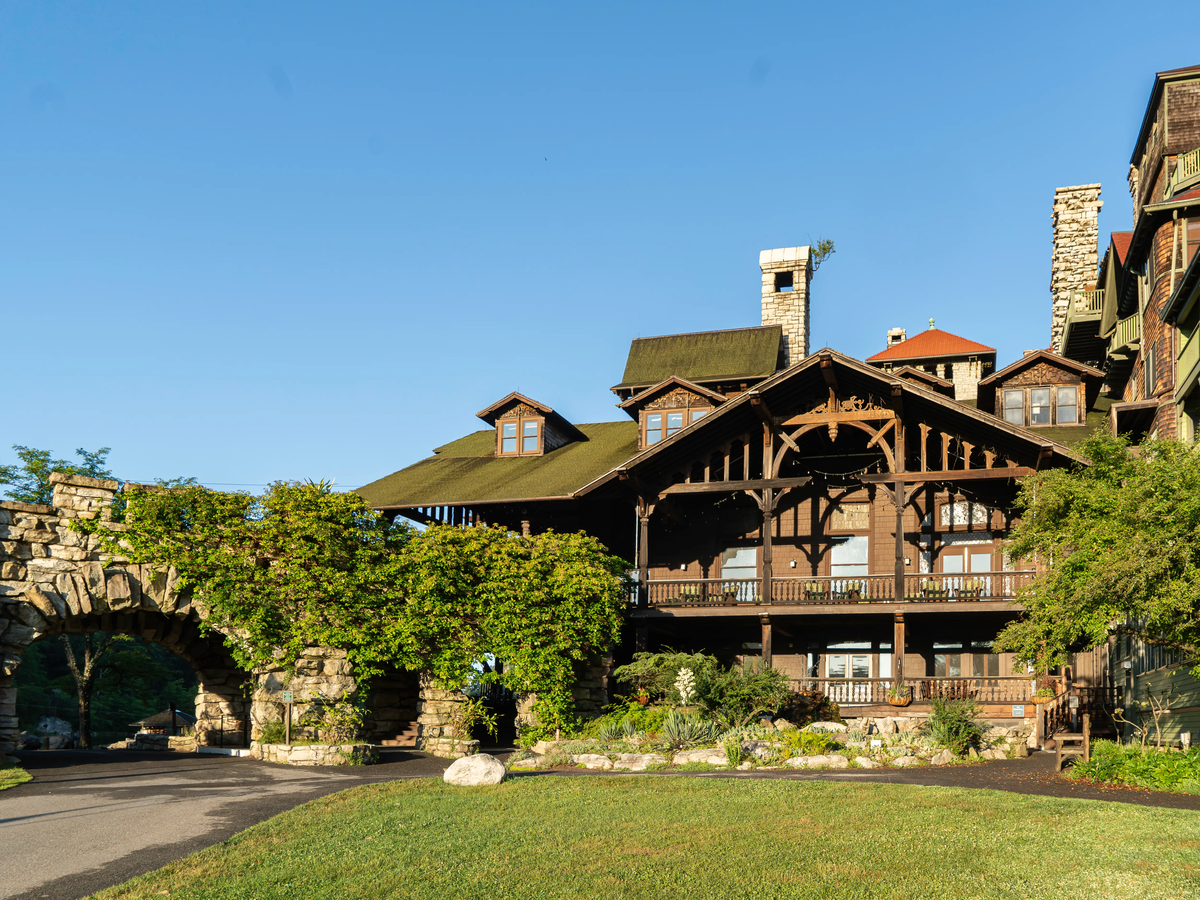Historic wooden resort building at Mohonk Mountain House with stone archways, balconies, chimneys, lawn, and trees under a clear blue sky.