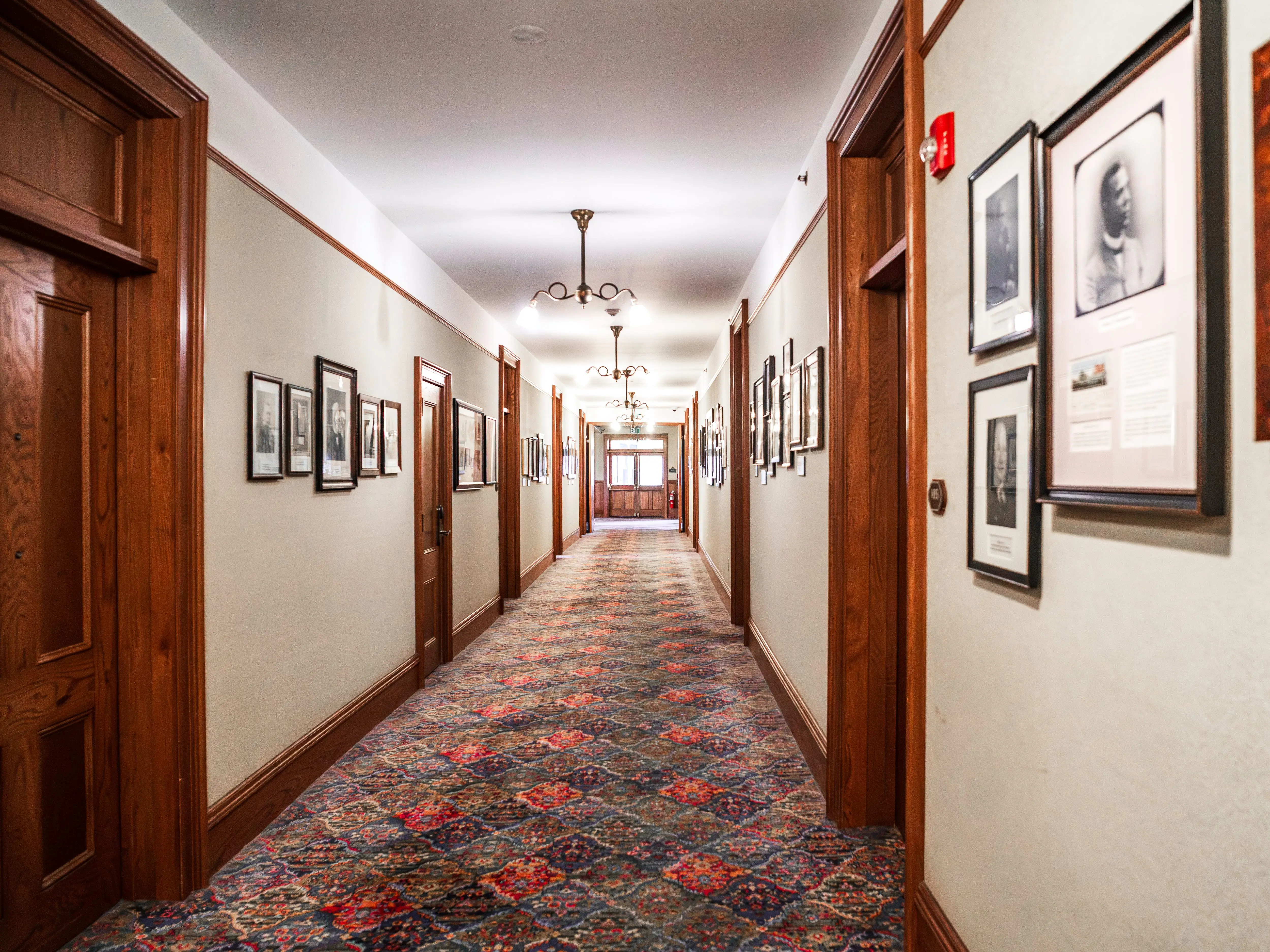 Long carpeted hallway with wood trim, doors, pendant lights, and framed photographs lining both walls.