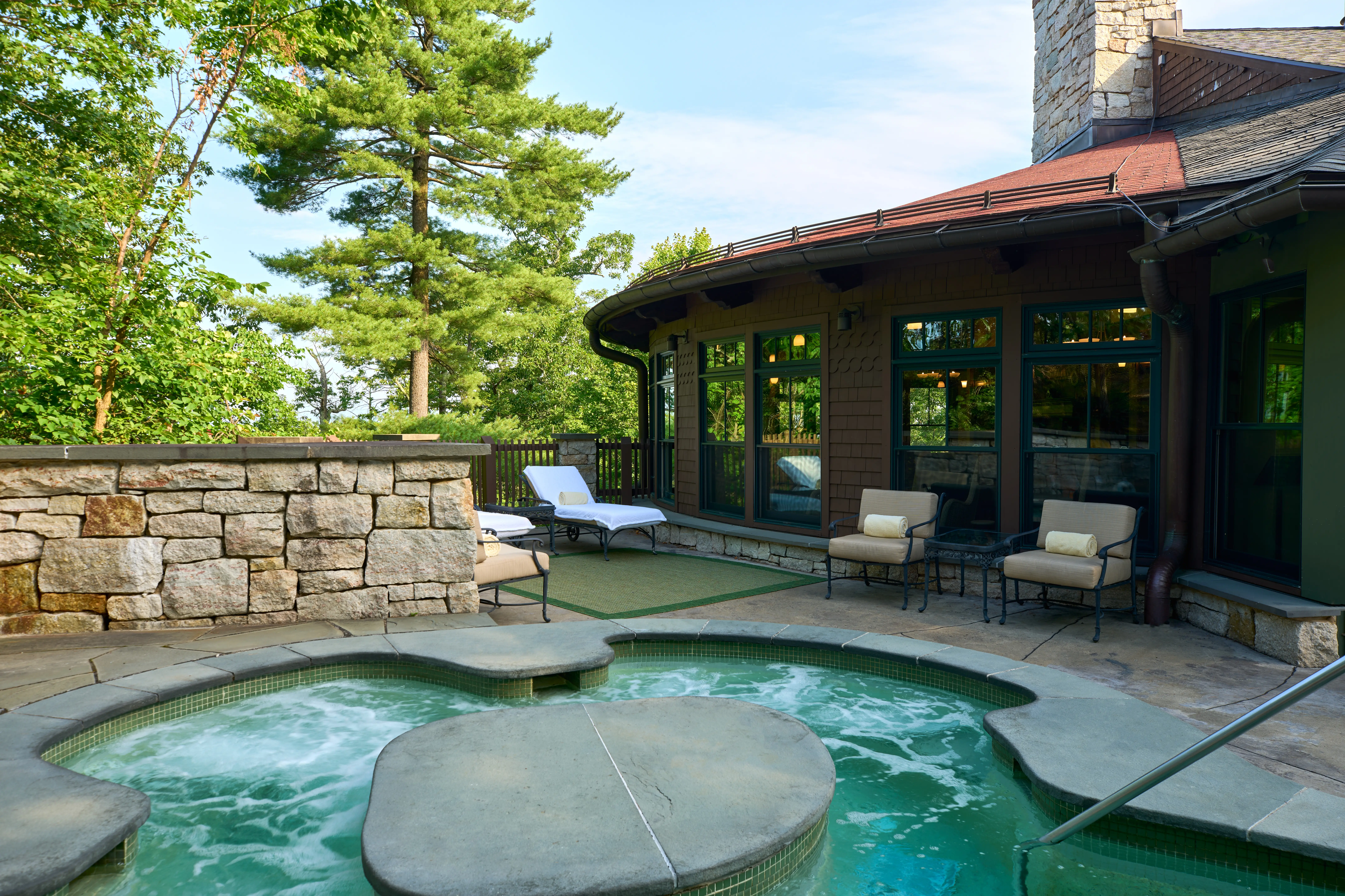 Outdoor hot tub patio with stone walls, lounge seating, and a rustic lodge building surrounded by trees.