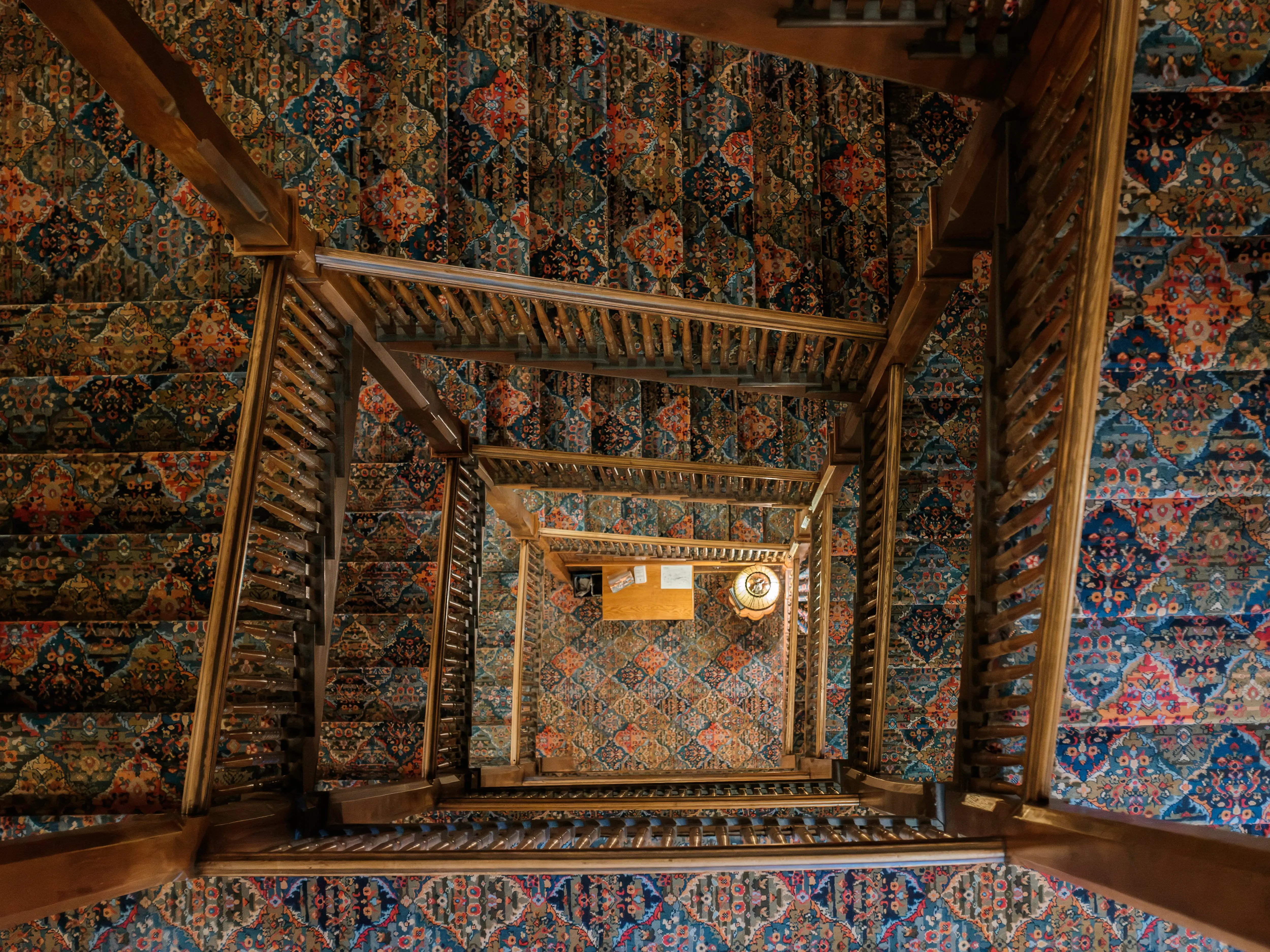 Ornate wooden stairwell viewed from above, surrounded by richly patterned blue, red, and orange carpeted surfaces.