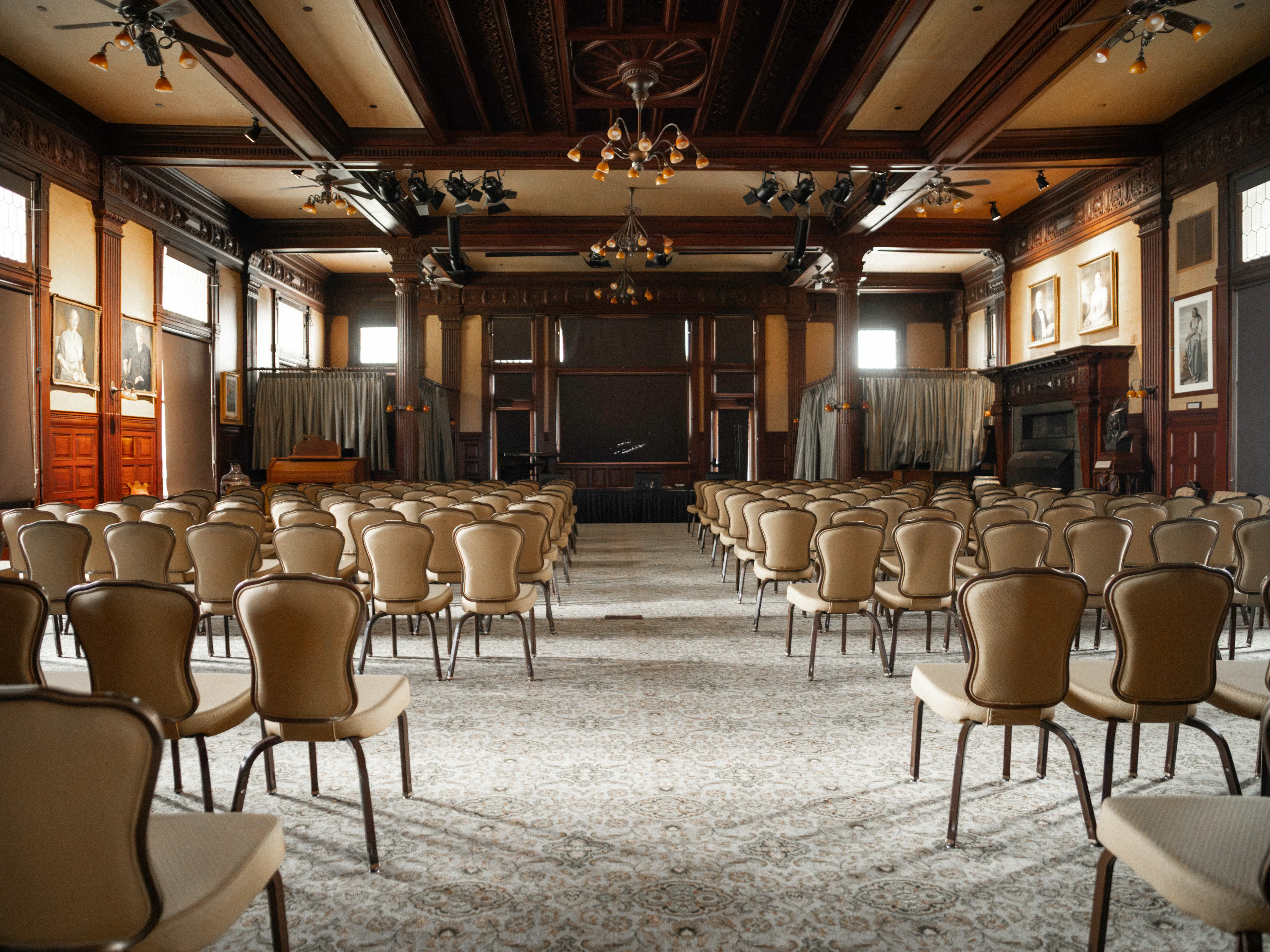 Rows of empty chairs face a small stage in an ornate wood-paneled hall with chandeliers and portraits.