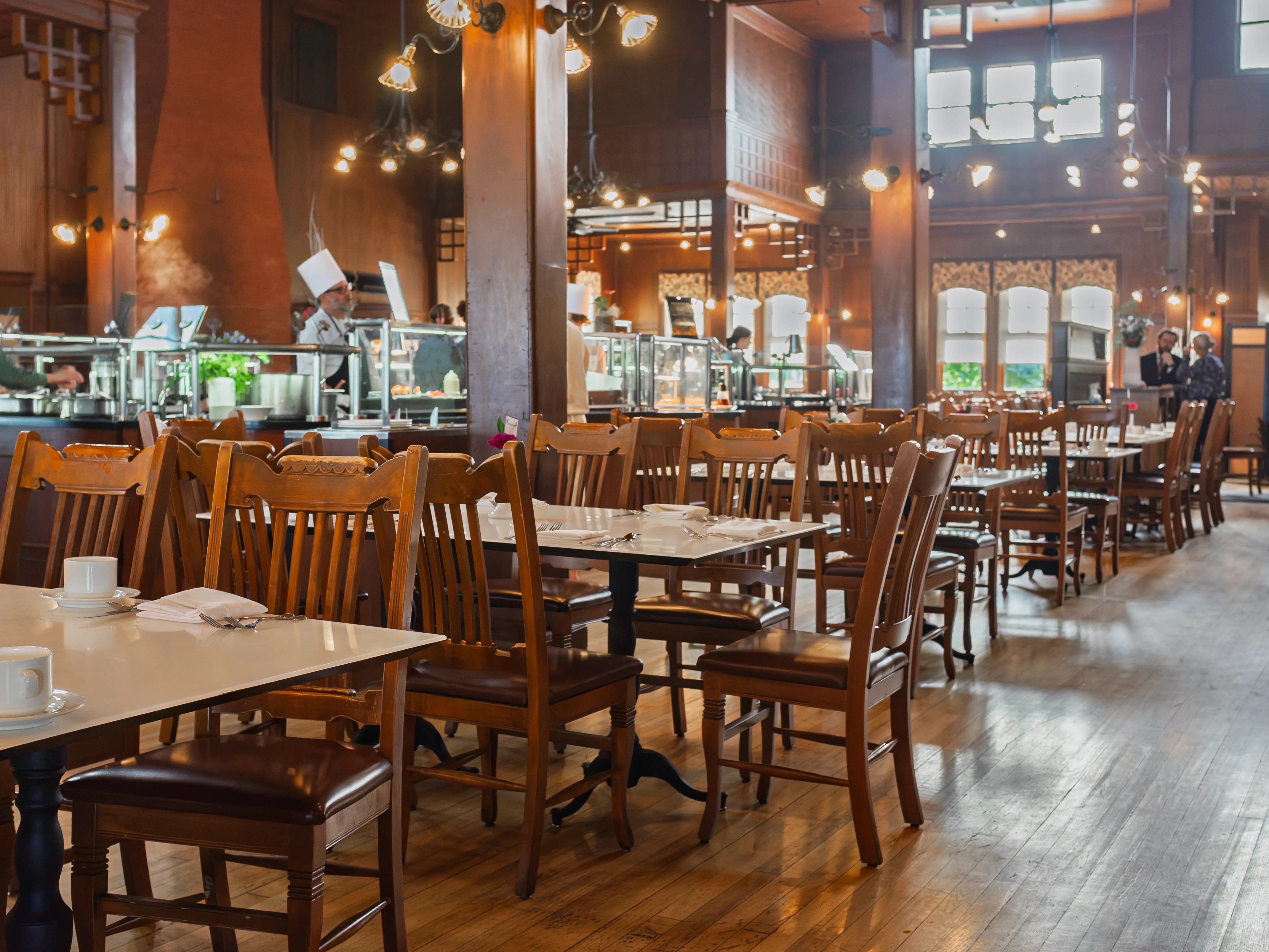 Wooden chairs and set tables fill a warmly lit restaurant dining room with chefs working at a buffet station.