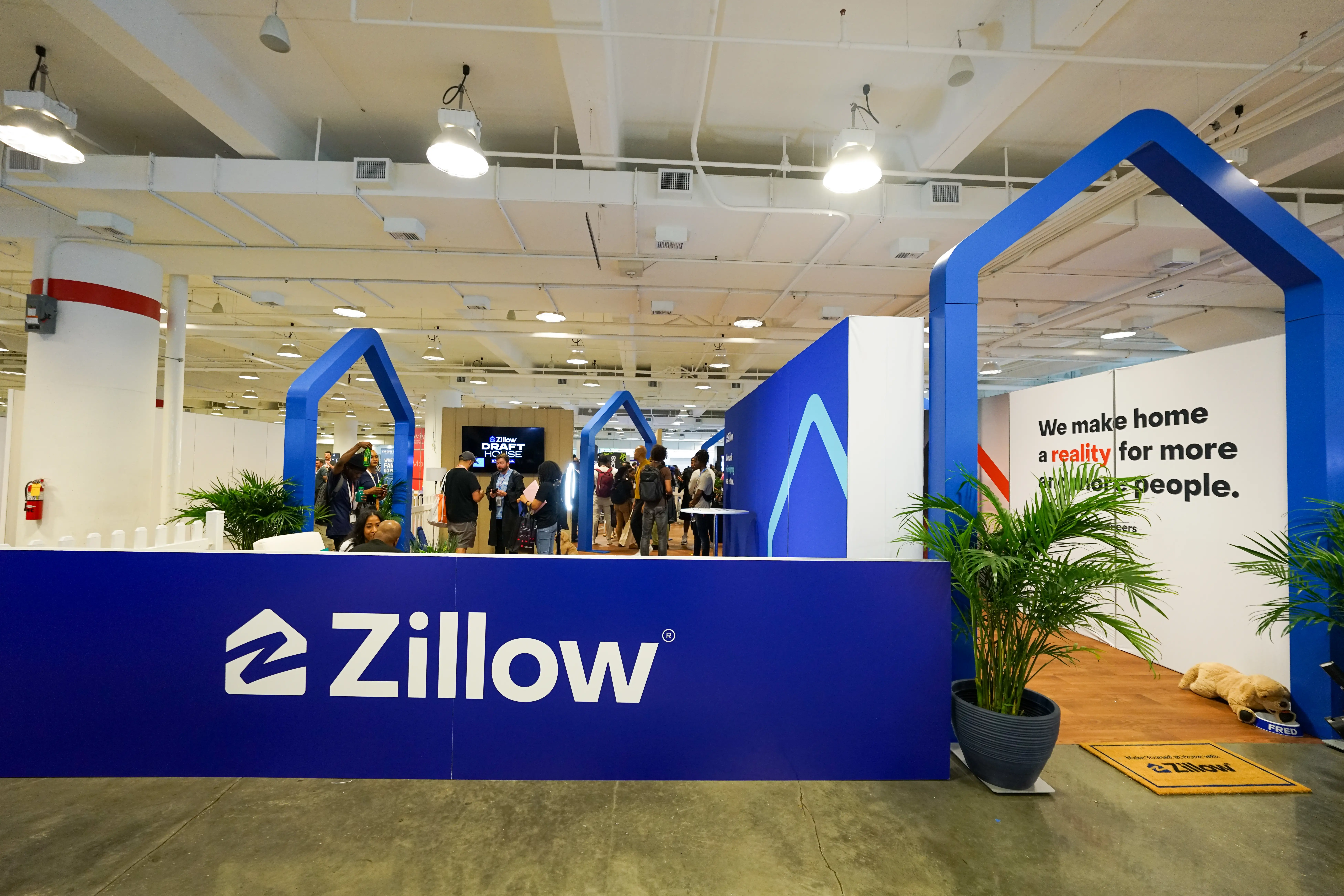 Zillow branded exhibition booth with blue house-shaped displays and visitors inside a large indoor event space.