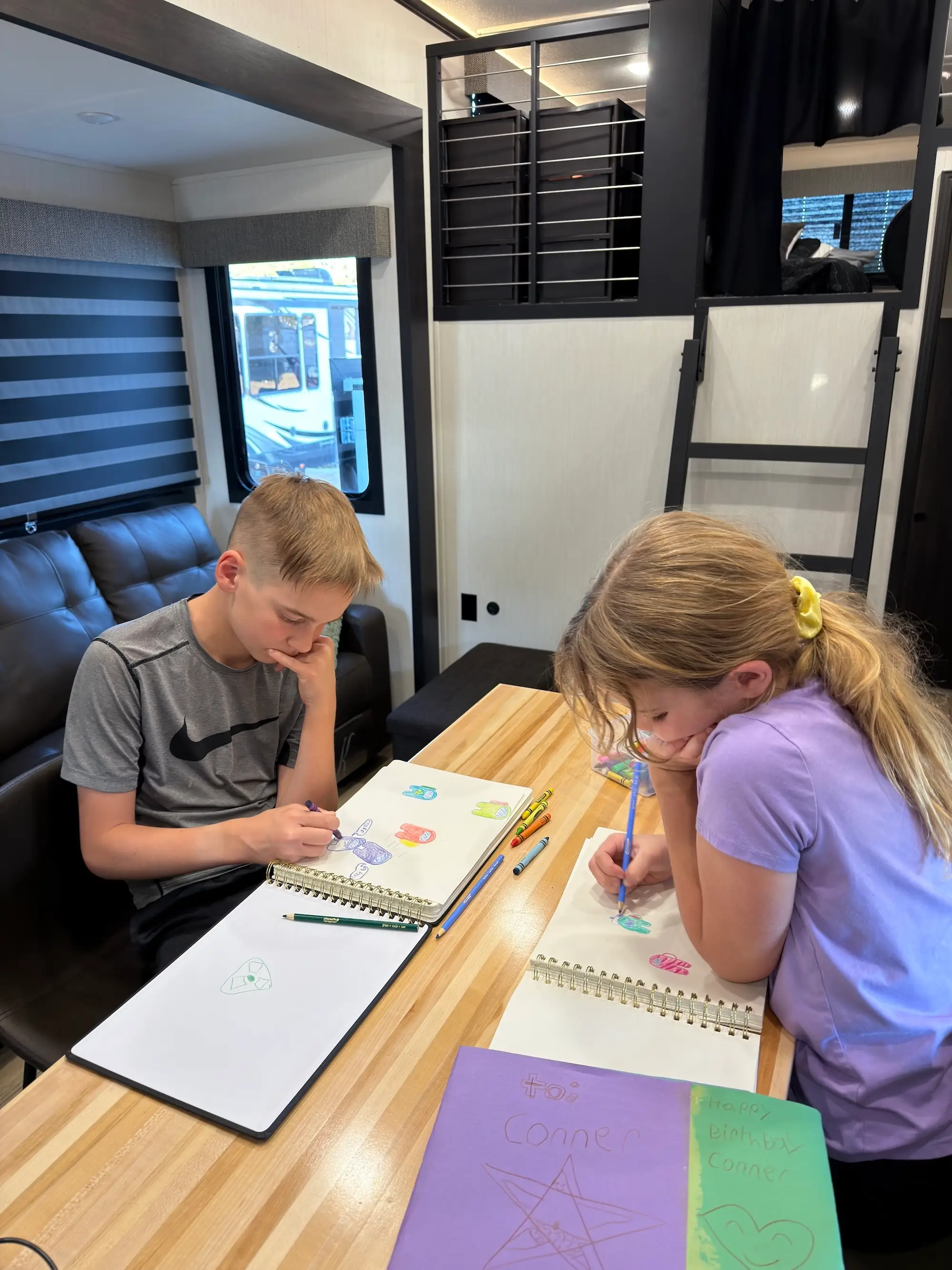 Two kids draw in spiral sketchbooks at a table inside a camper with a lofted bed and window.