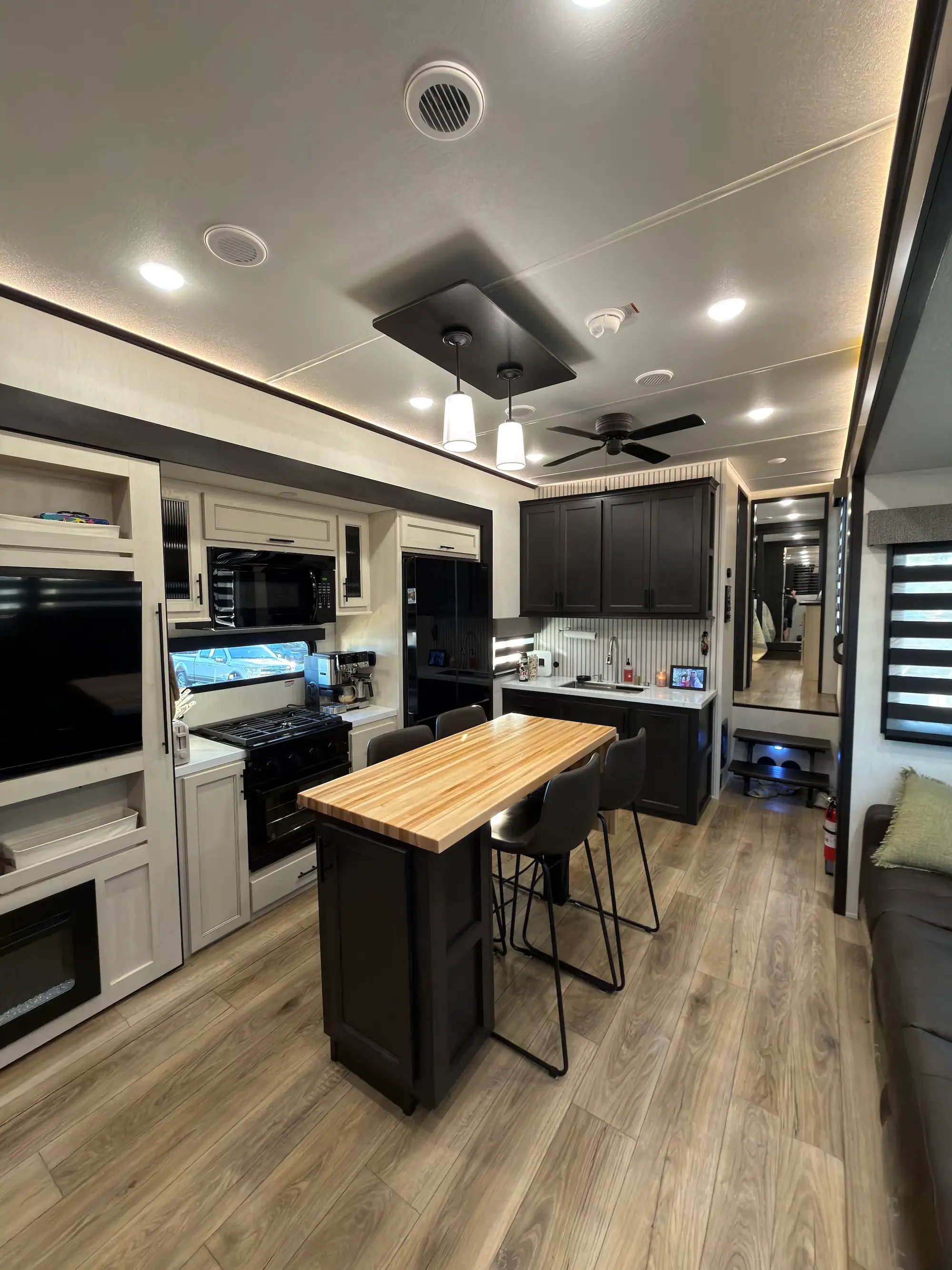 Modern RV kitchen and living area with island seating, appliances, ceiling lights, fan, sofa, and wood-look flooring.
