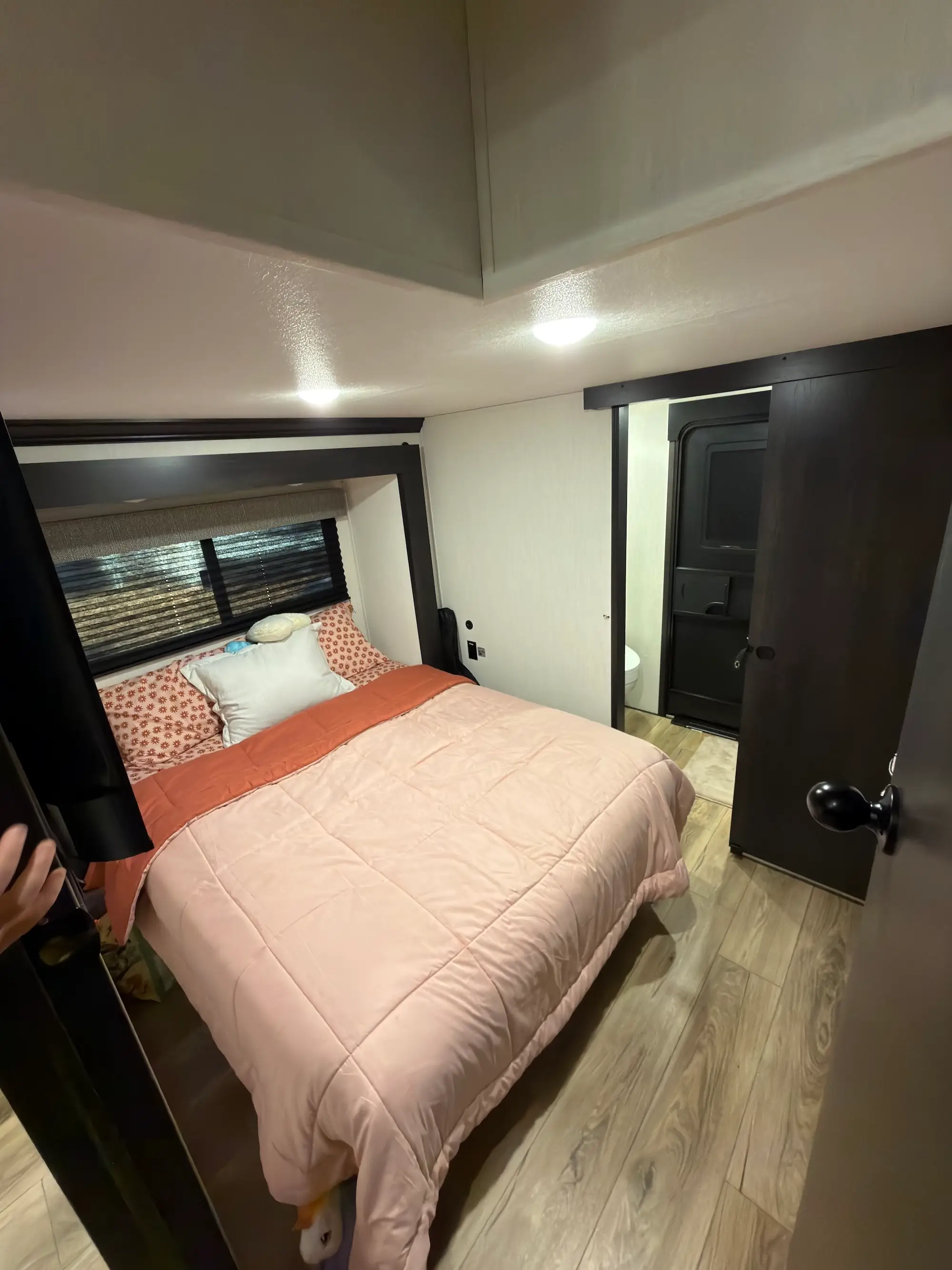 Compact bedroom with a made bed, window blinds, overhead lights, wood-look flooring, and a sliding bathroom doorway.