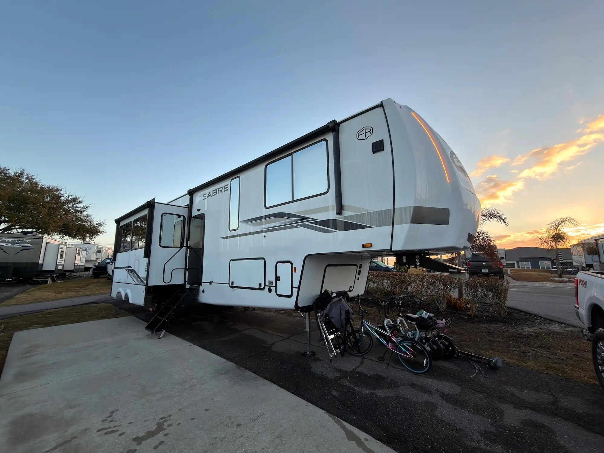 White SABRE fifth-wheel RV parked at an RV park at sunset with bicycles nearby.
