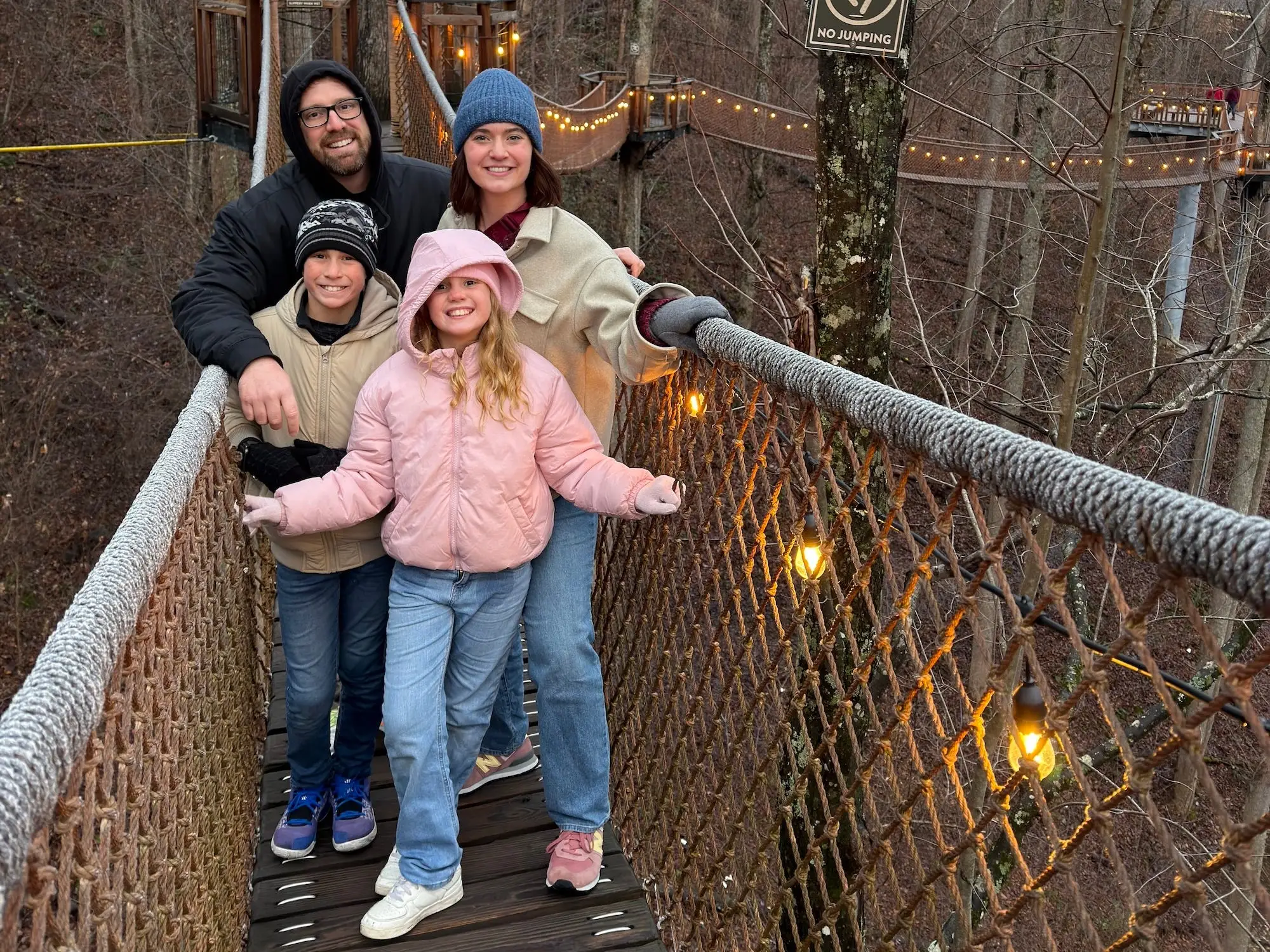 Two adults and two children pose on a rope suspension bridge with string lights in a leafless forest.