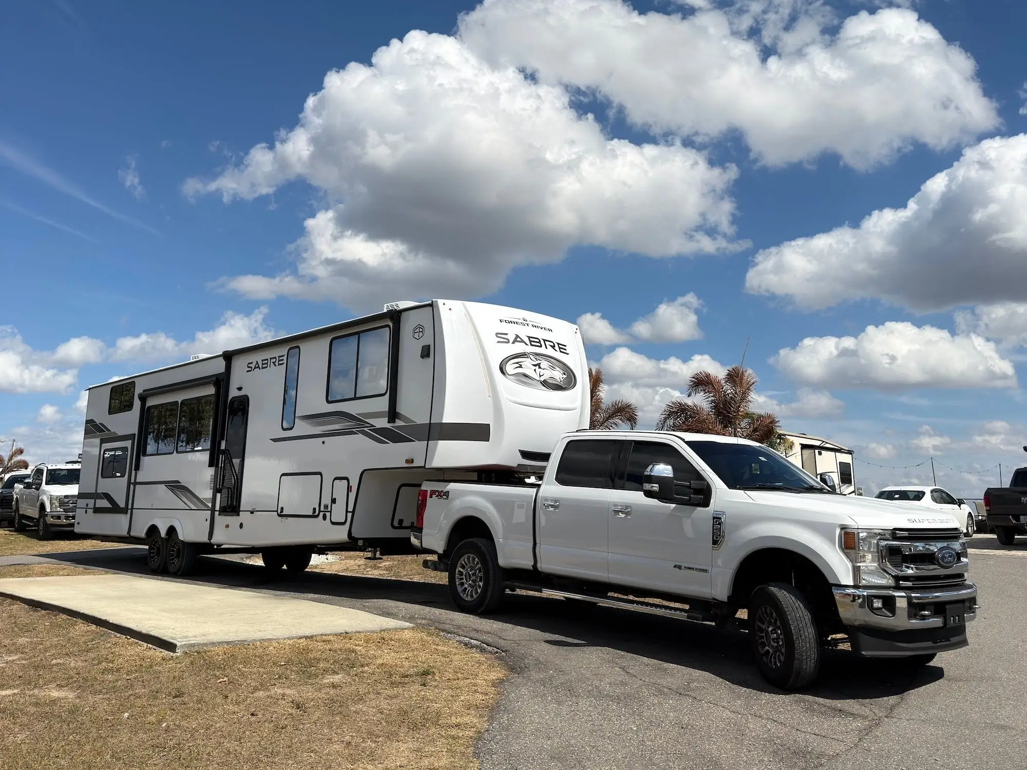 White Ford Super Duty pickup tows a SABRE fifth-wheel RV through a paved lot under a cloudy blue sky.
