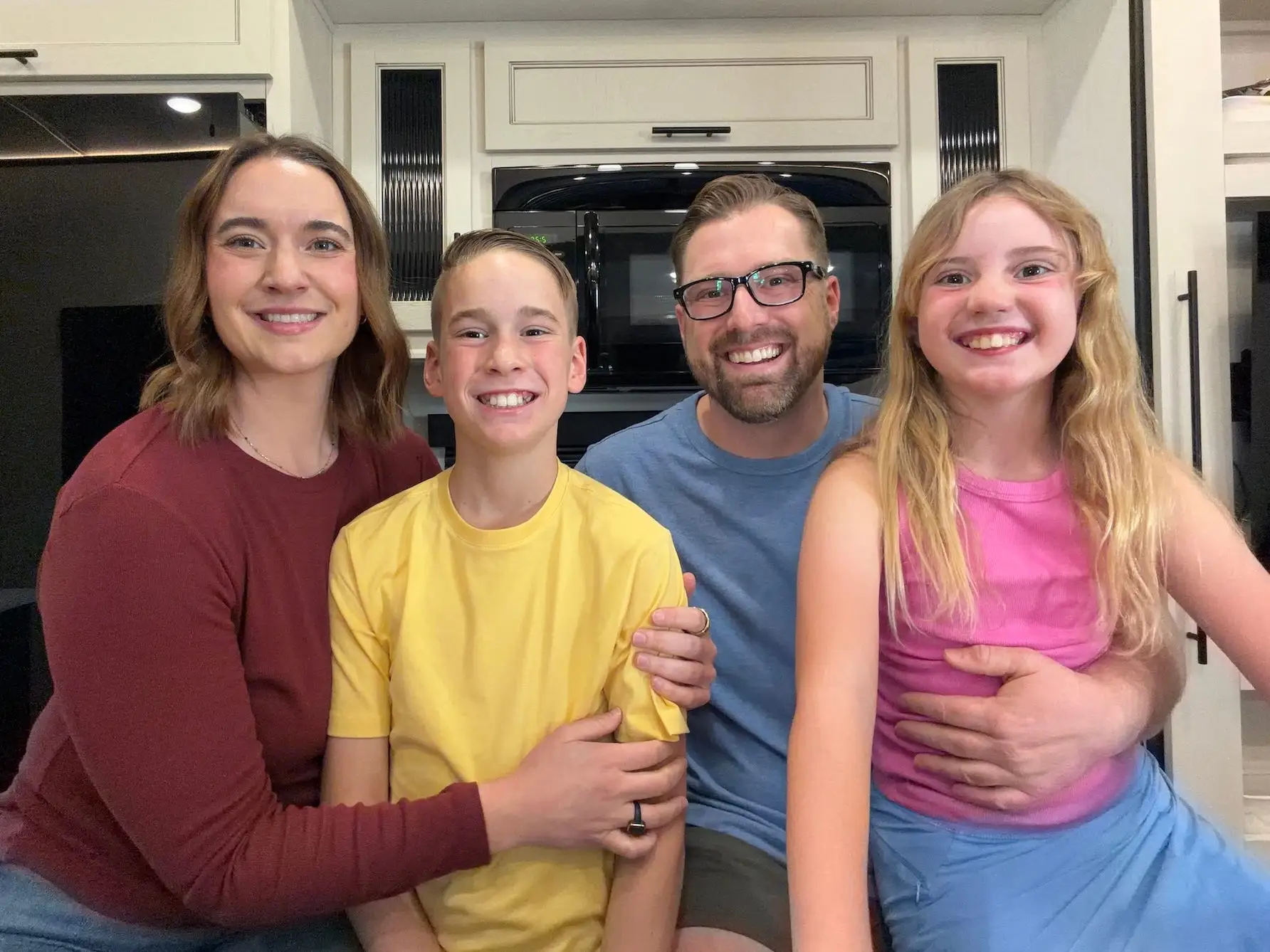Four smiling people sit close together in a compact kitchen interior with light cabinets and dark appliances.