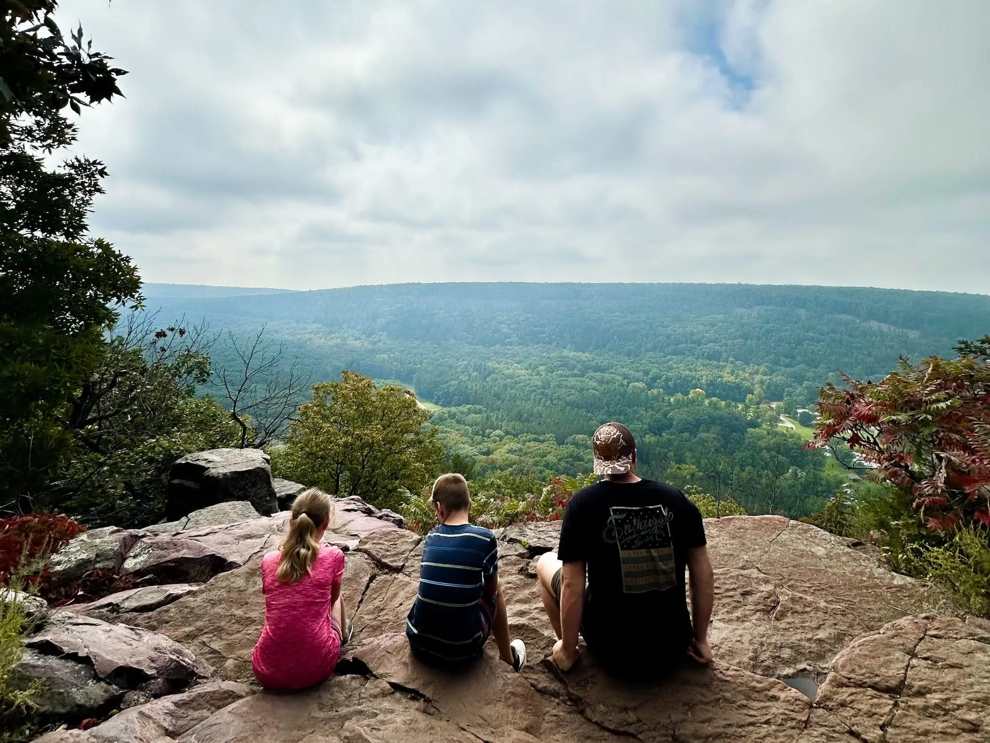 Three hikers sit on a rocky overlook facing a forested valley under a cloudy sky.