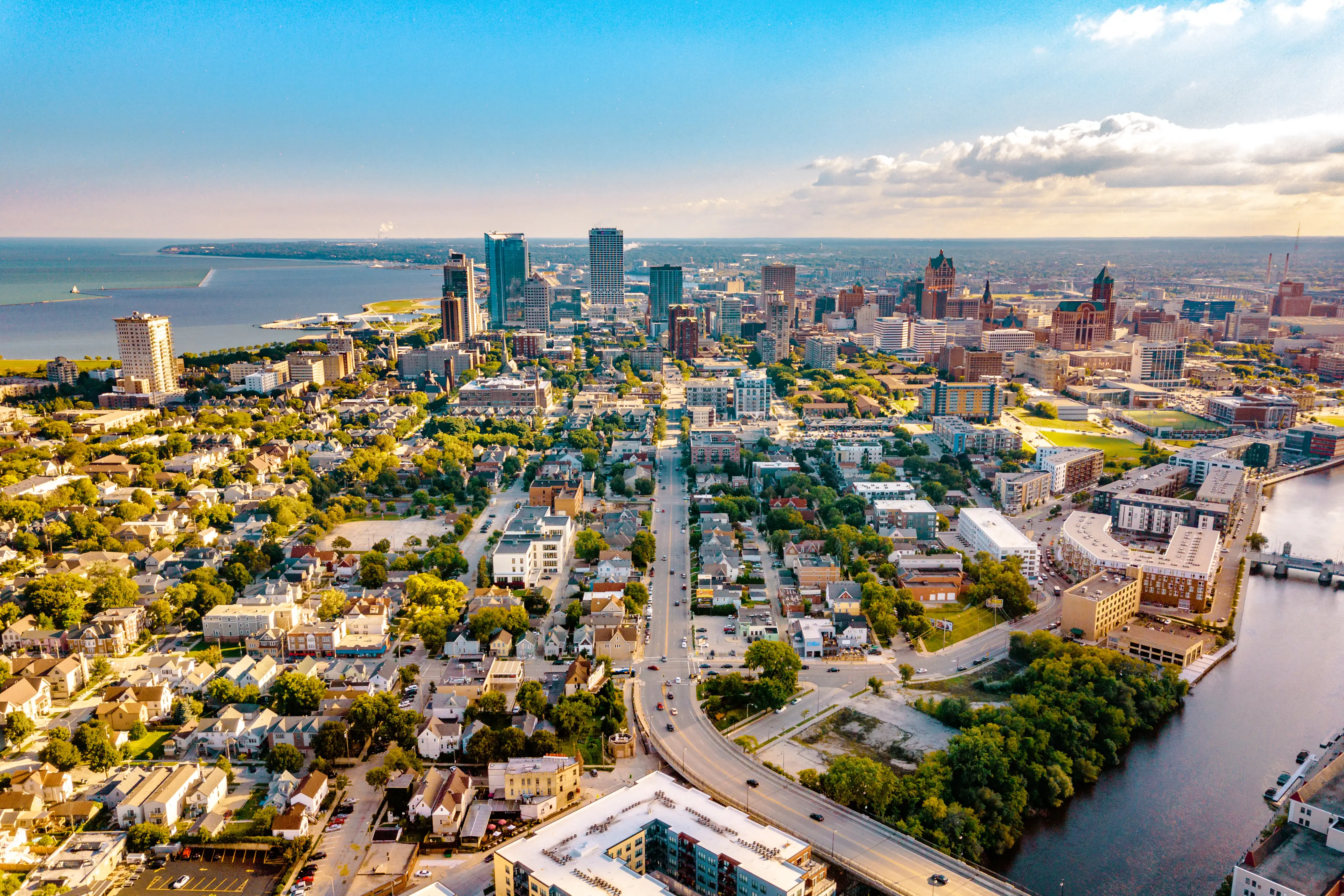 Aerial view of downtown Milwaukee skyline with surrounding neighborhoods, Lake Michigan, and the Milwaukee River at sunset.