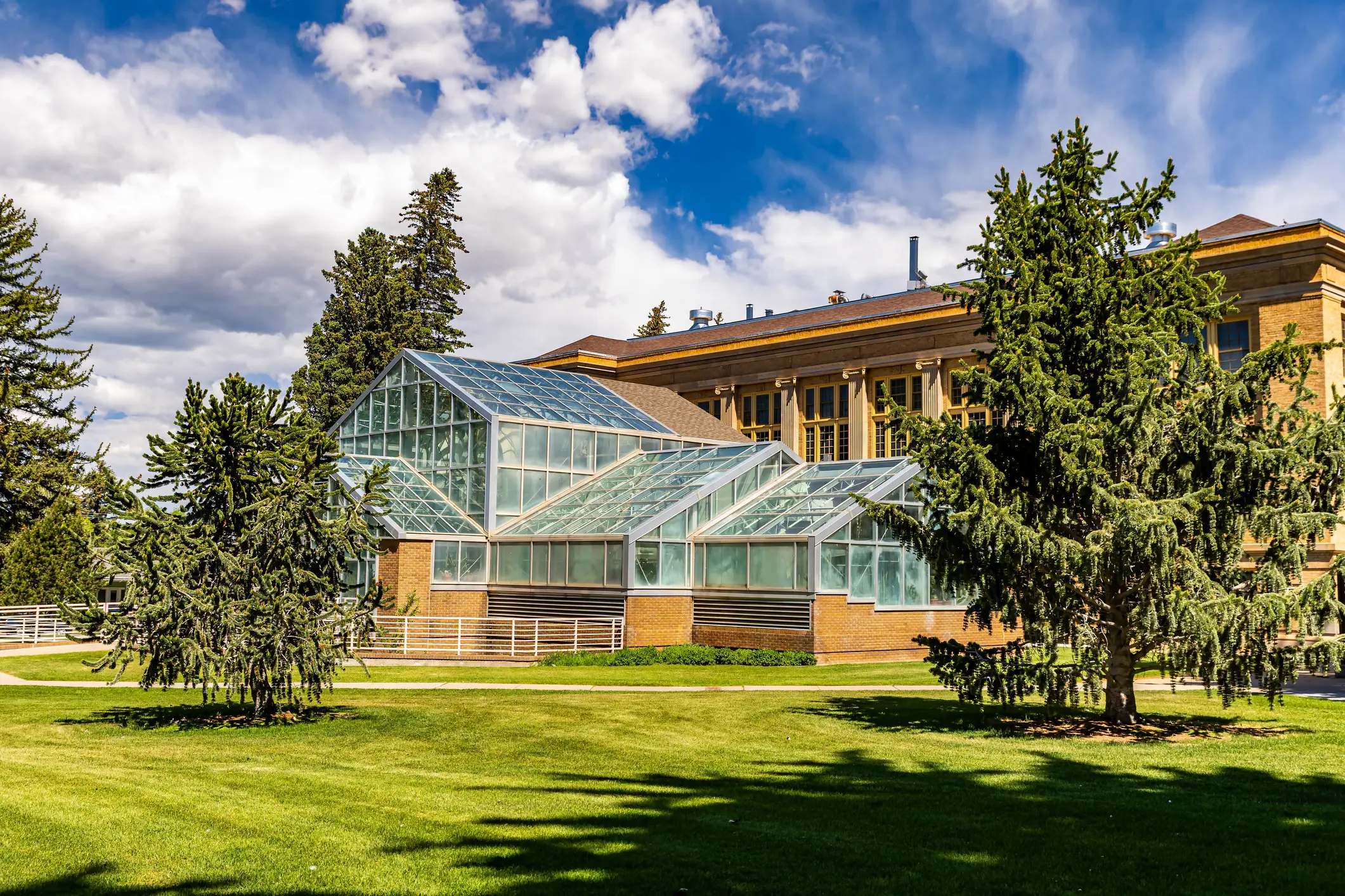 Glass greenhouse structure stands beside a brick building on a landscaped lawn under a partly cloudy sky.
