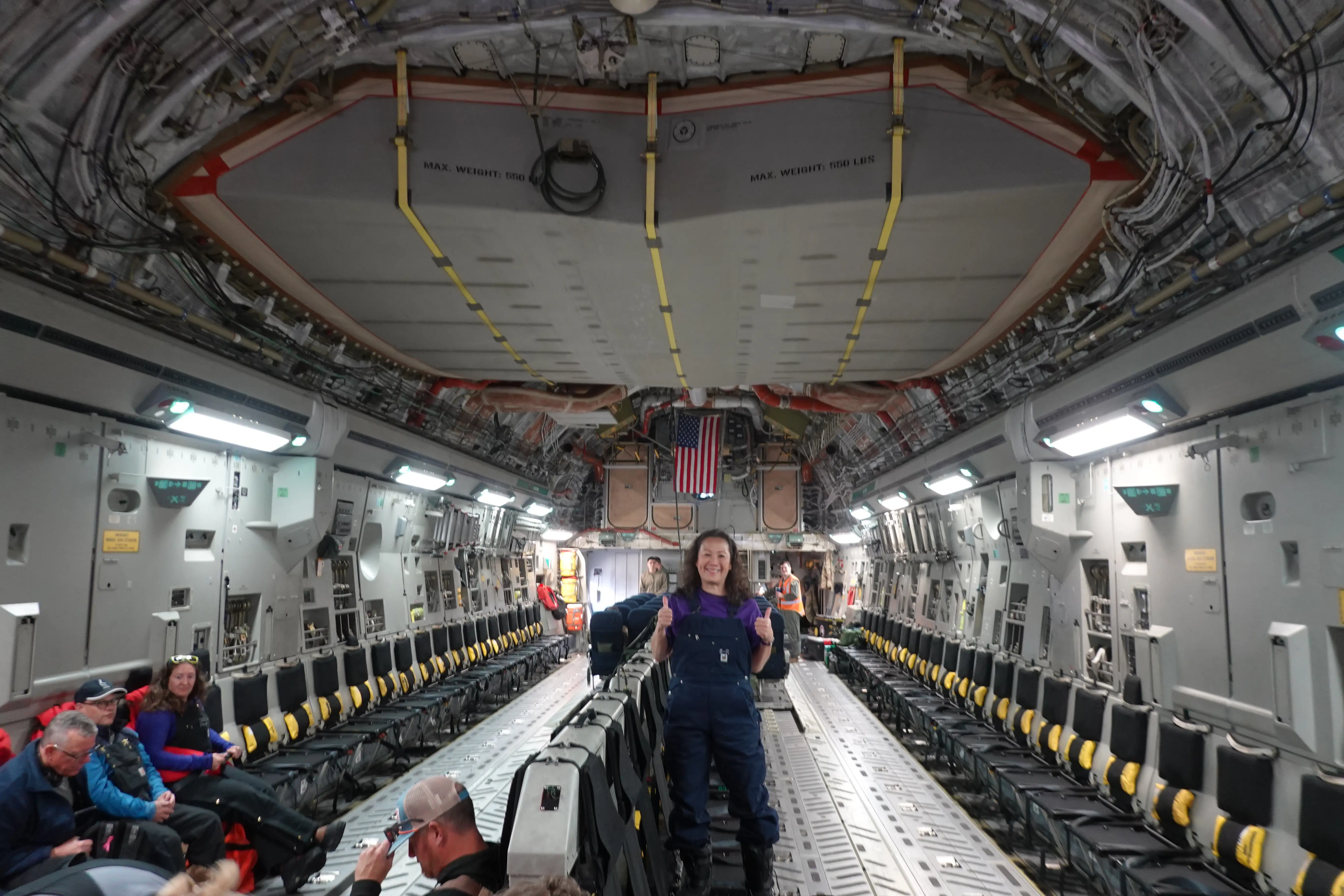 A person gives thumbs up inside a large aircraft cabin lined with fold-down seats and an American flag.