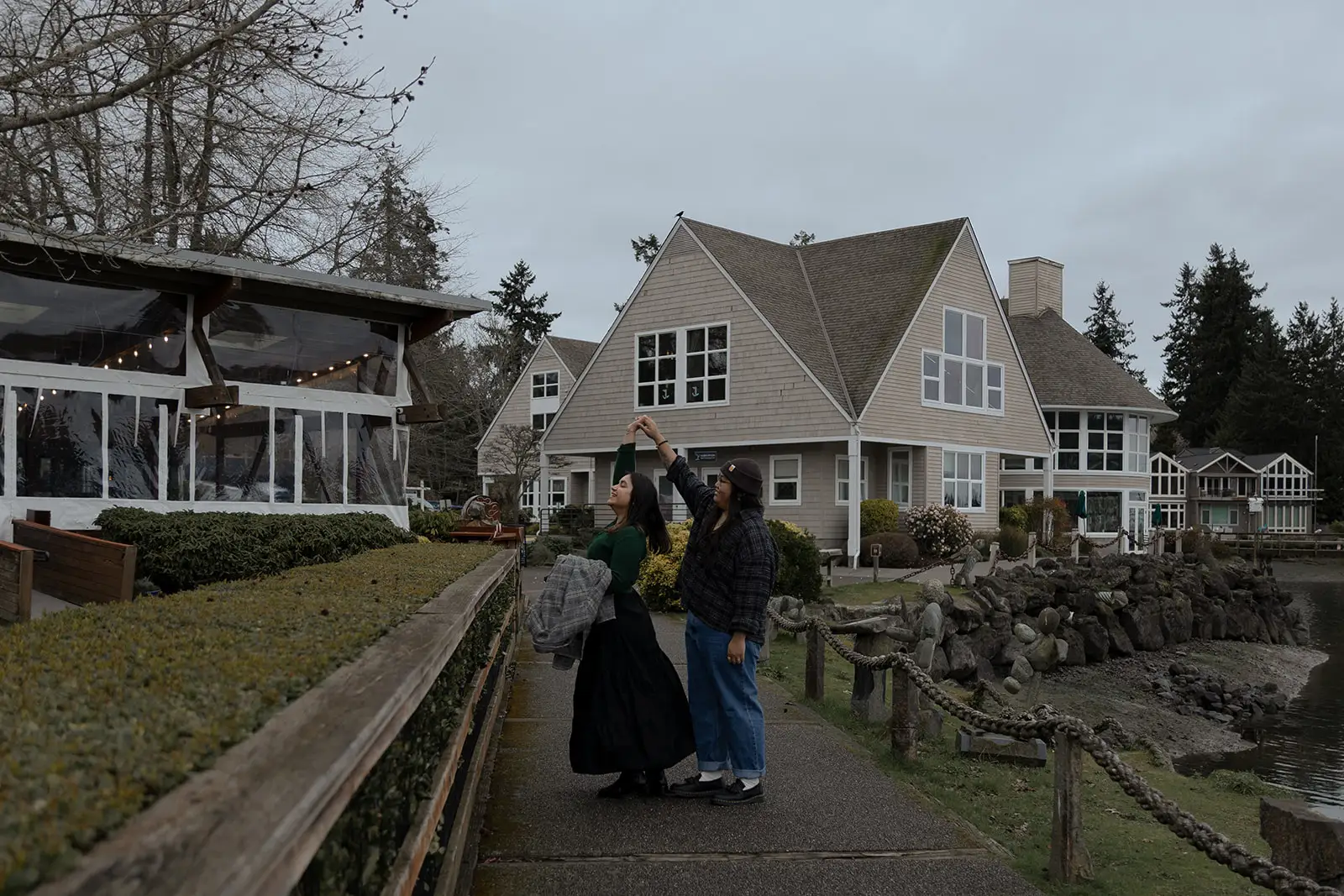 Two people hold hands on a waterfront path beside shingled buildings under an overcast sky.