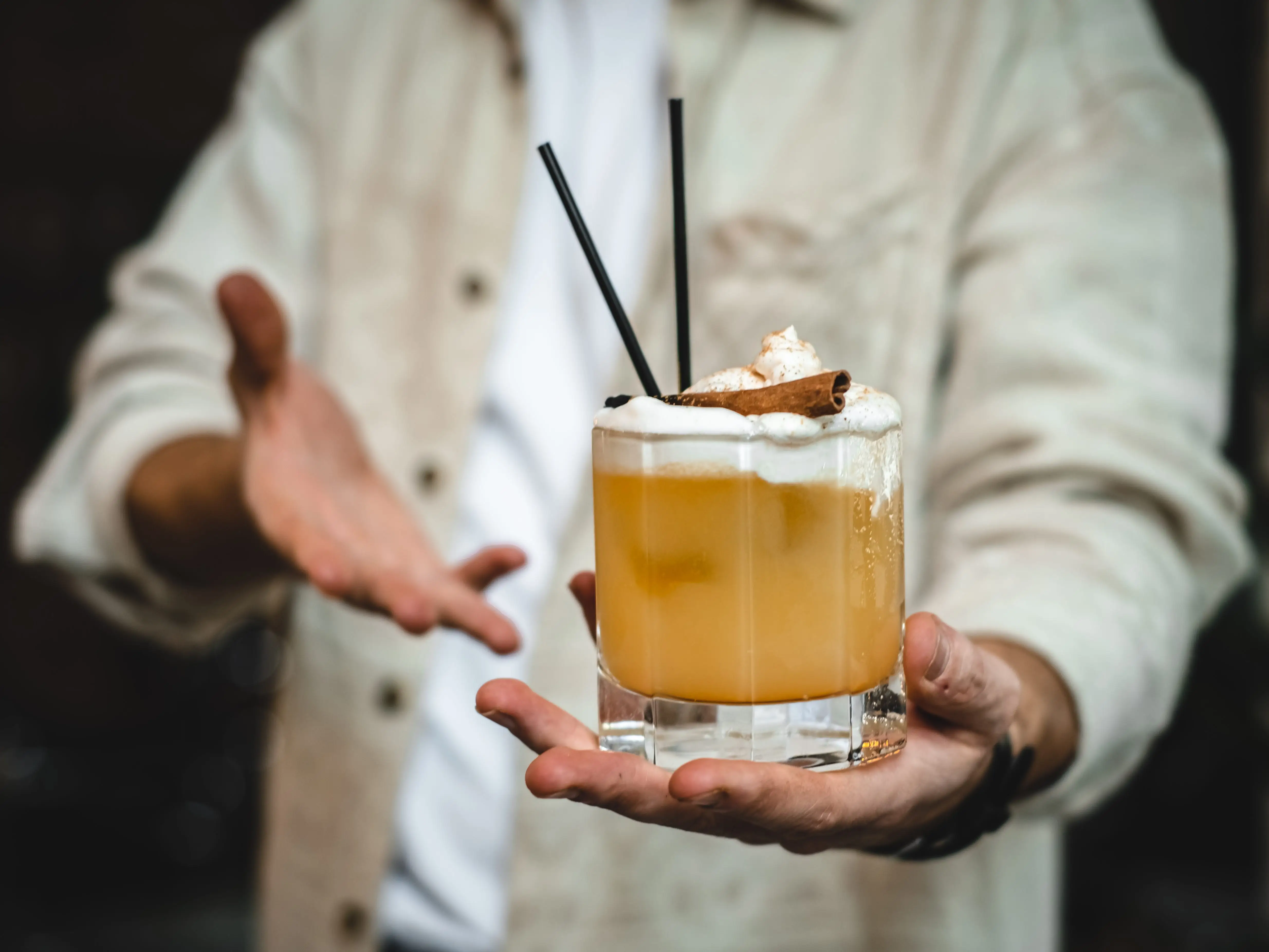 hand holding orange-yellow cocktail with foam and cinnamon stick on top