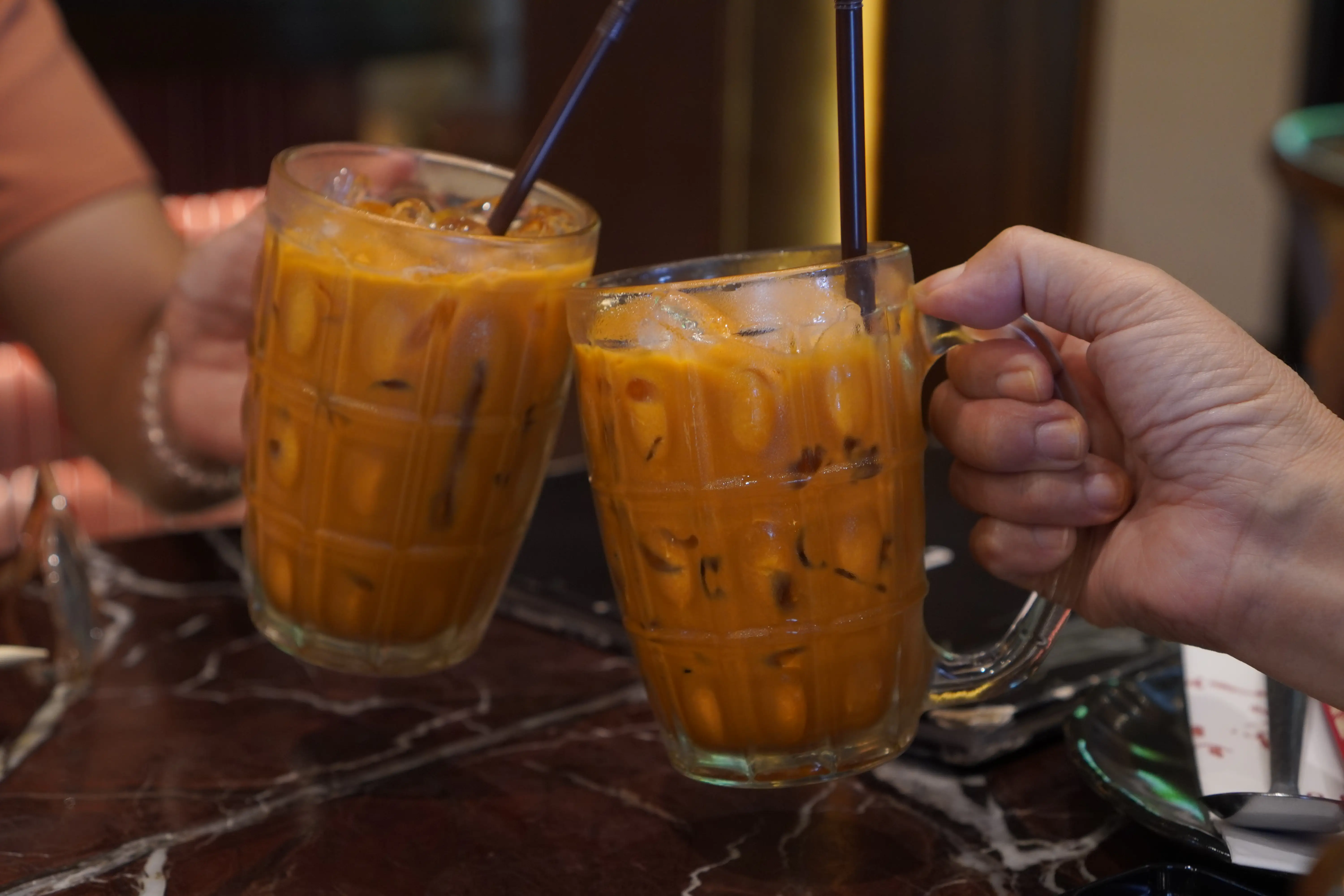 Two people toasting Thai tea-based drinks in glass mugs together