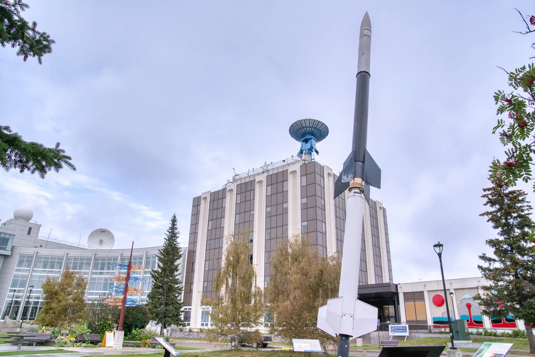 Tall rocket monument stands beside a multi-story building with antenna dishes in a landscaped outdoor plaza.