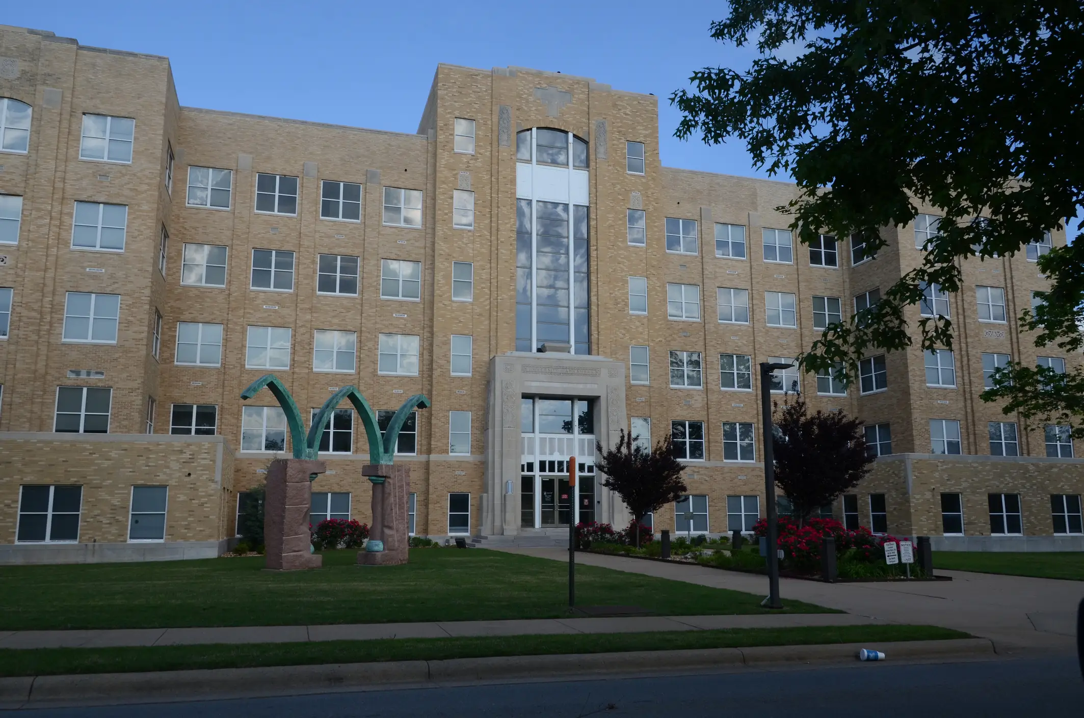 Large beige brick institutional building with rows of windows, a central entrance, lawn, sidewalk, and outdoor sculpture.