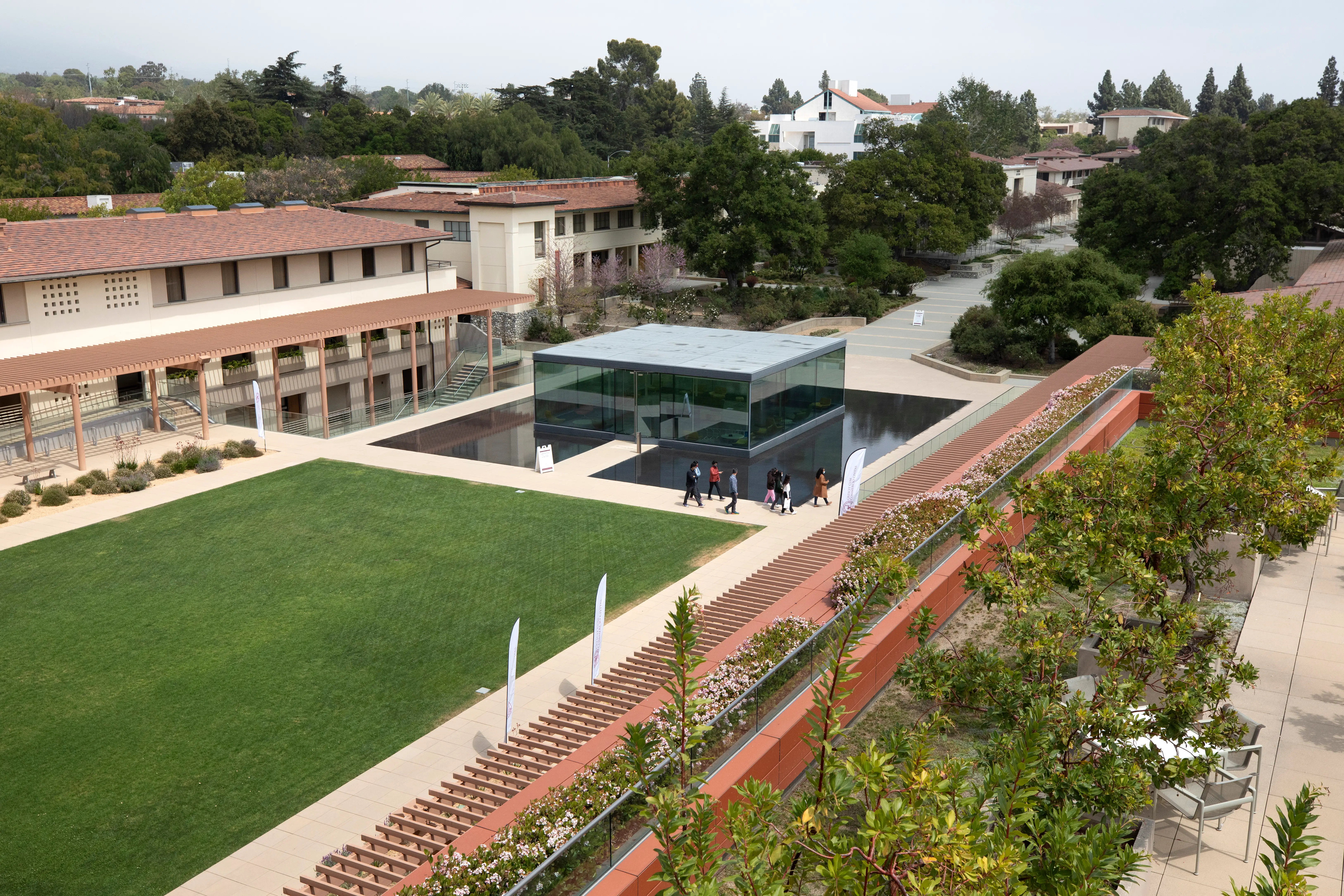 Courtyard campus with lawn, tiled-roof buildings, a glass pavilion, reflecting pool, trees, and people walking on paths.