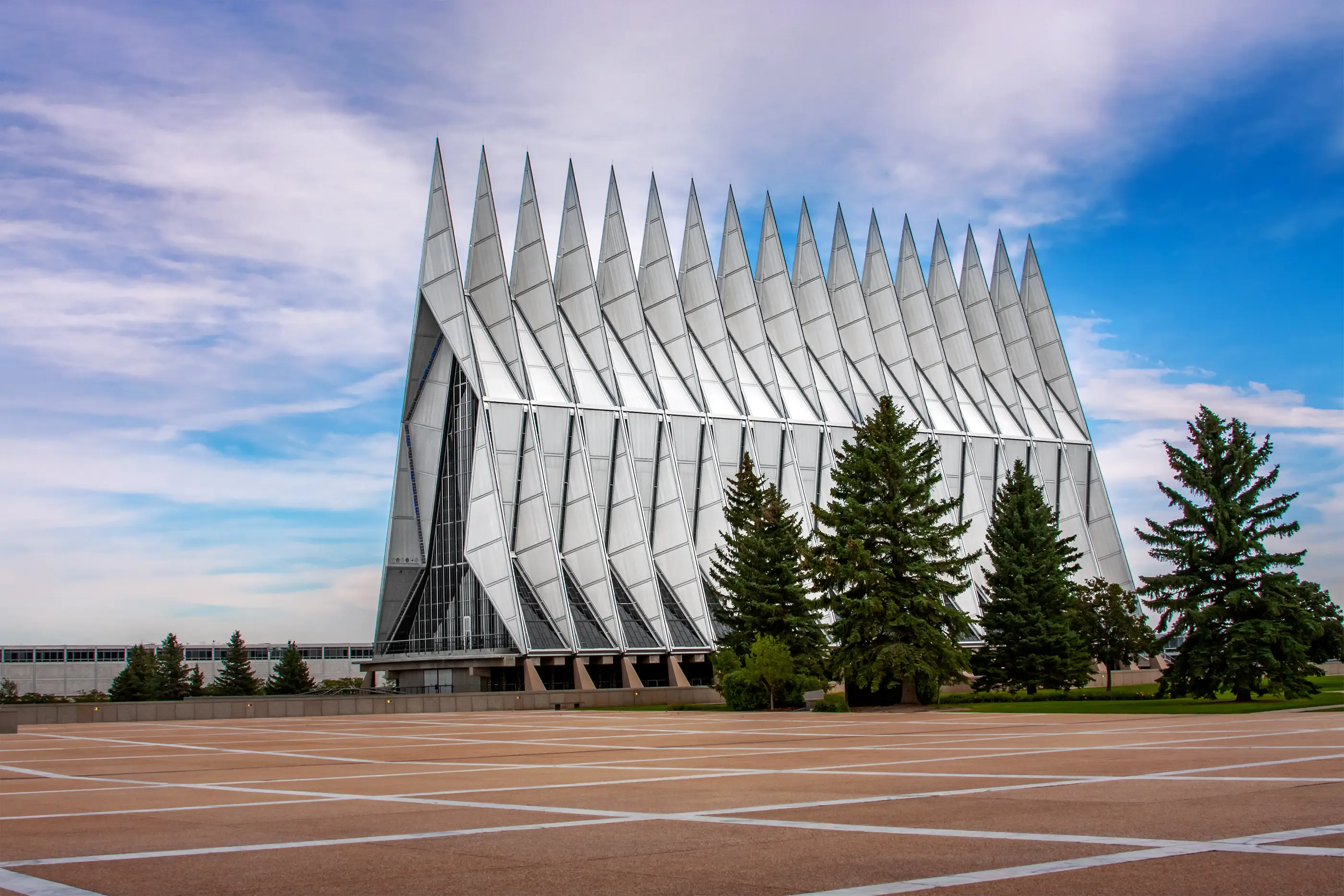 United States Air Force Academy Cadet Chapel stands behind evergreen trees and a patterned plaza under a partly cloudy sky.