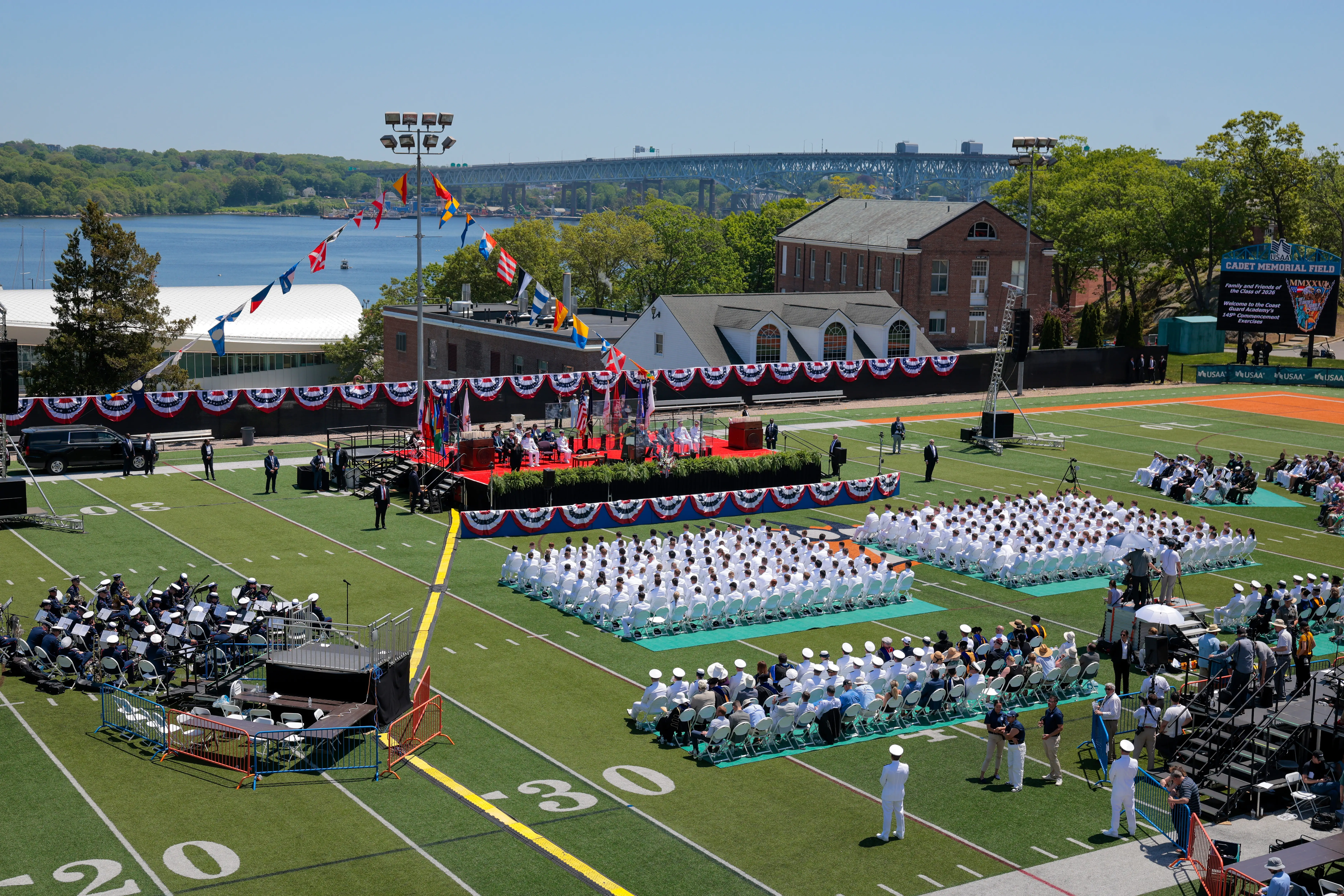 Uniformed graduates sit on a football field during an outdoor commencement ceremony by the water.