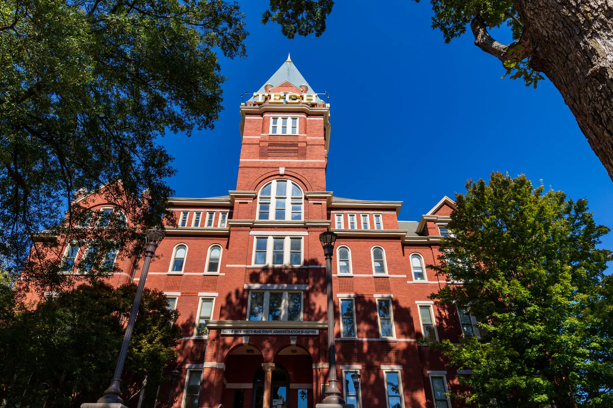 Red brick campus tower with TECH sign rises behind trees under a clear blue sky.