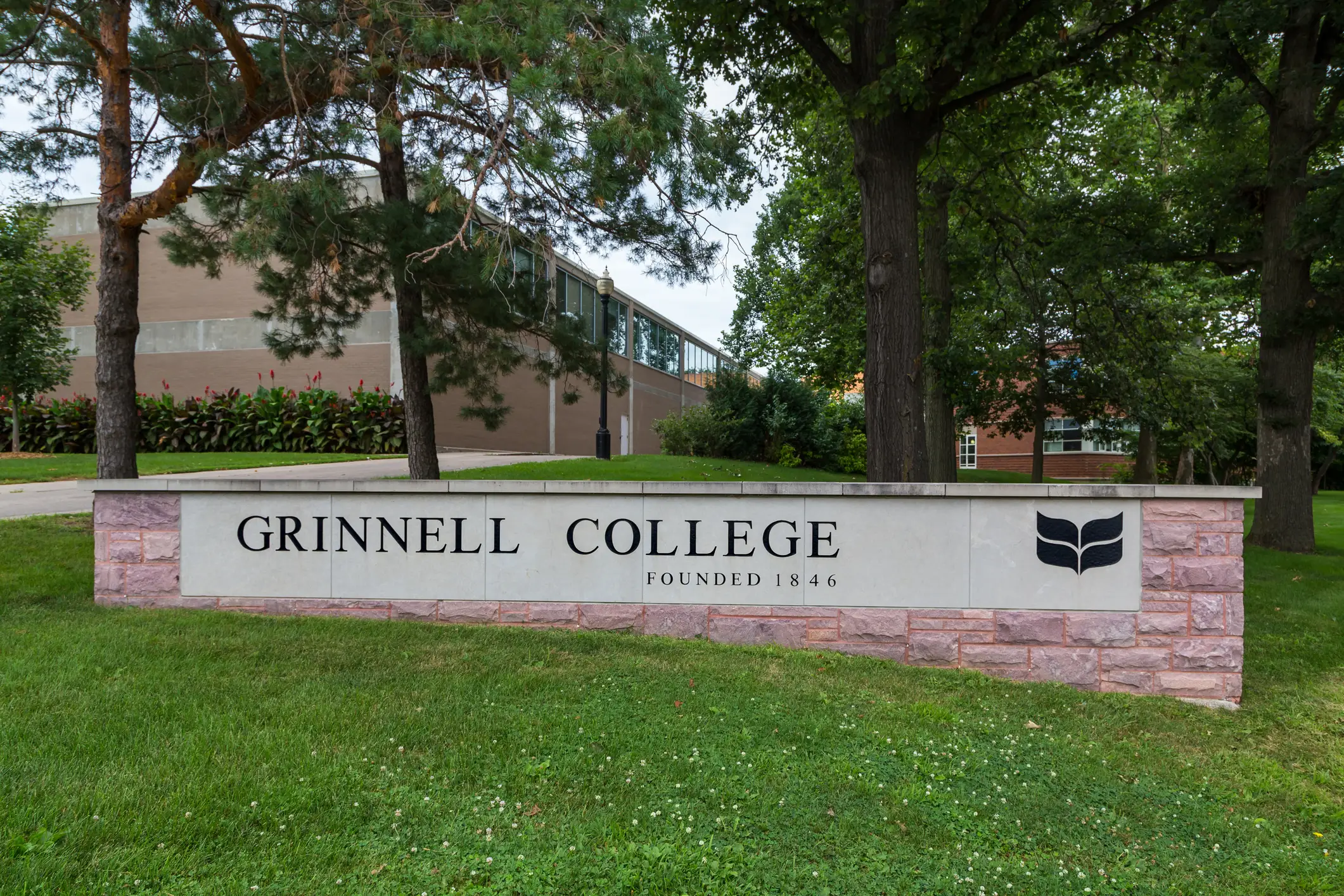 Grinnell College entrance sign stands on a grassy campus lawn surrounded by trees and brick buildings.