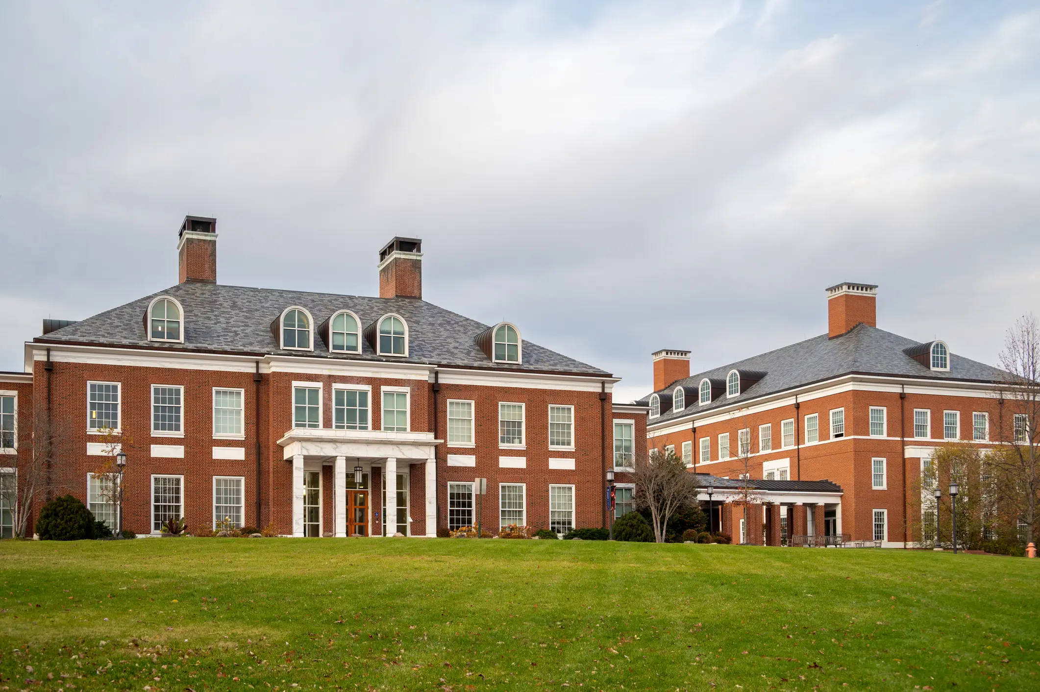 Red-brick institutional buildings with white-trimmed windows stand behind a broad green lawn under a cloudy sky.