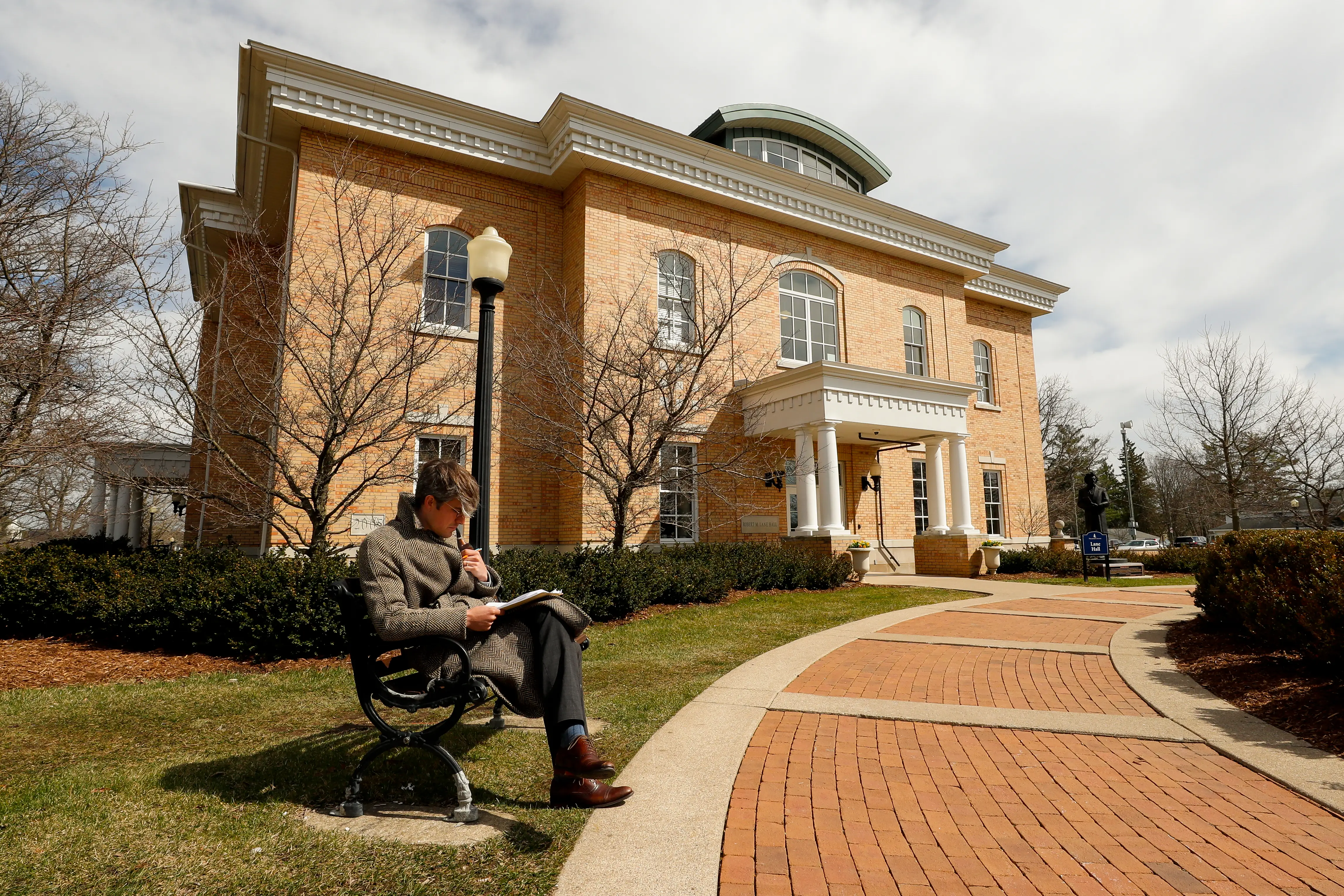 A person sits on a bench with a notebook outside a large brick building with columns and arched windows.