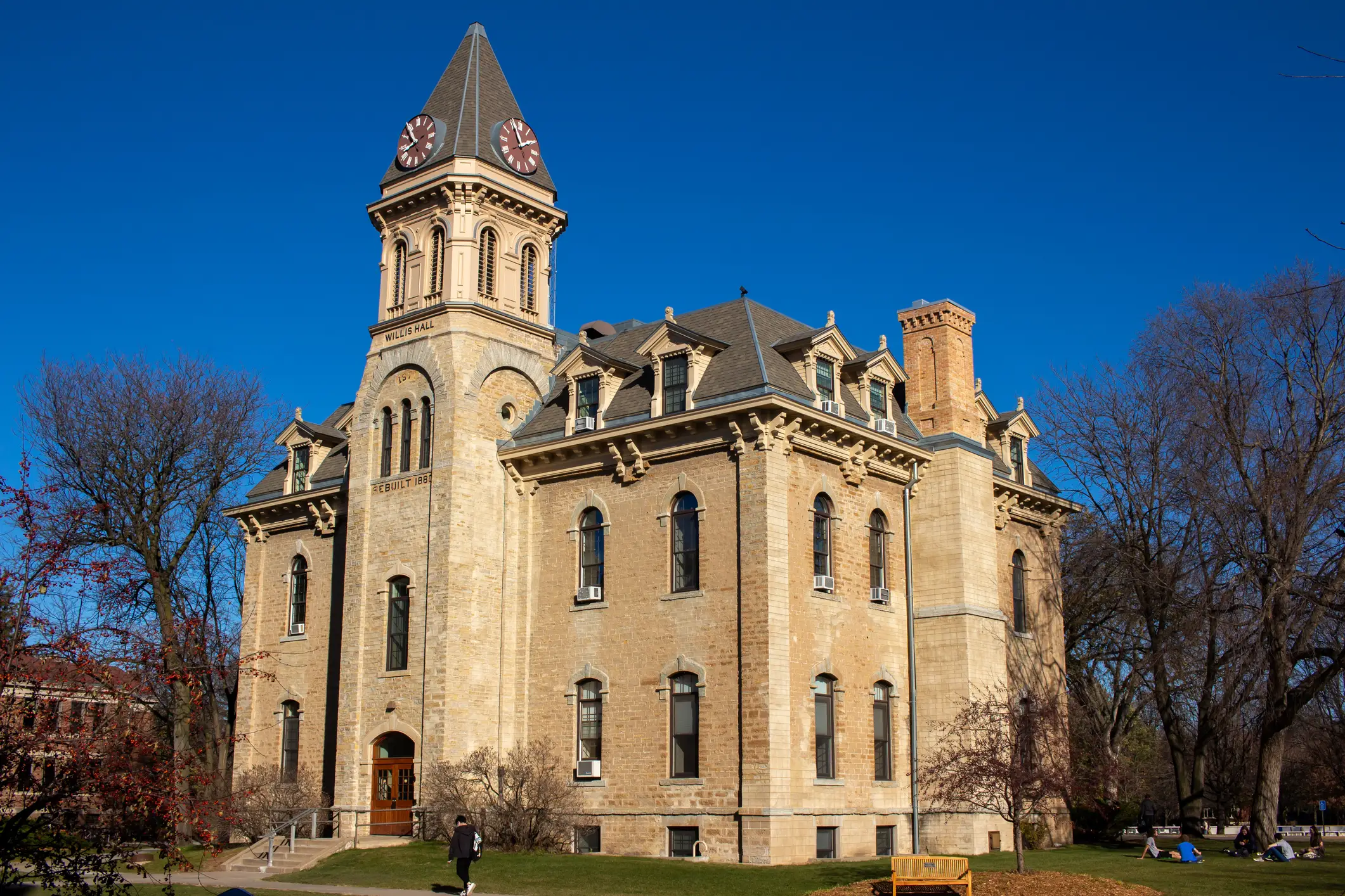 Historic stone Wills Hall with a clock tower stands on a campus lawn under a clear blue sky.