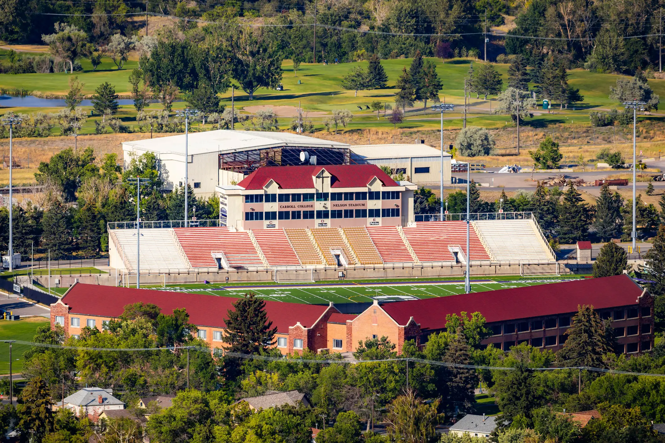 Aerial view of Carroll College Nelson Stadium with football field, bleachers, campus buildings, and surrounding trees.
