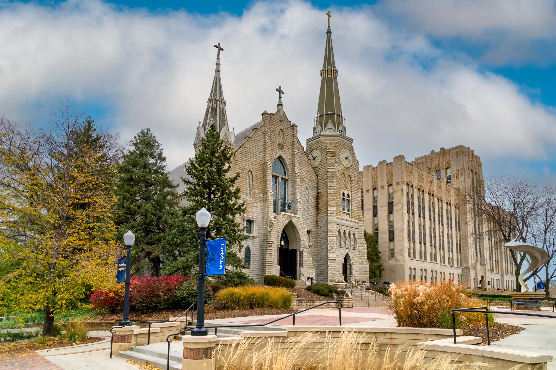 Stone church with twin spires and crosses stands beside Creighton banners on a landscaped campus plaza.