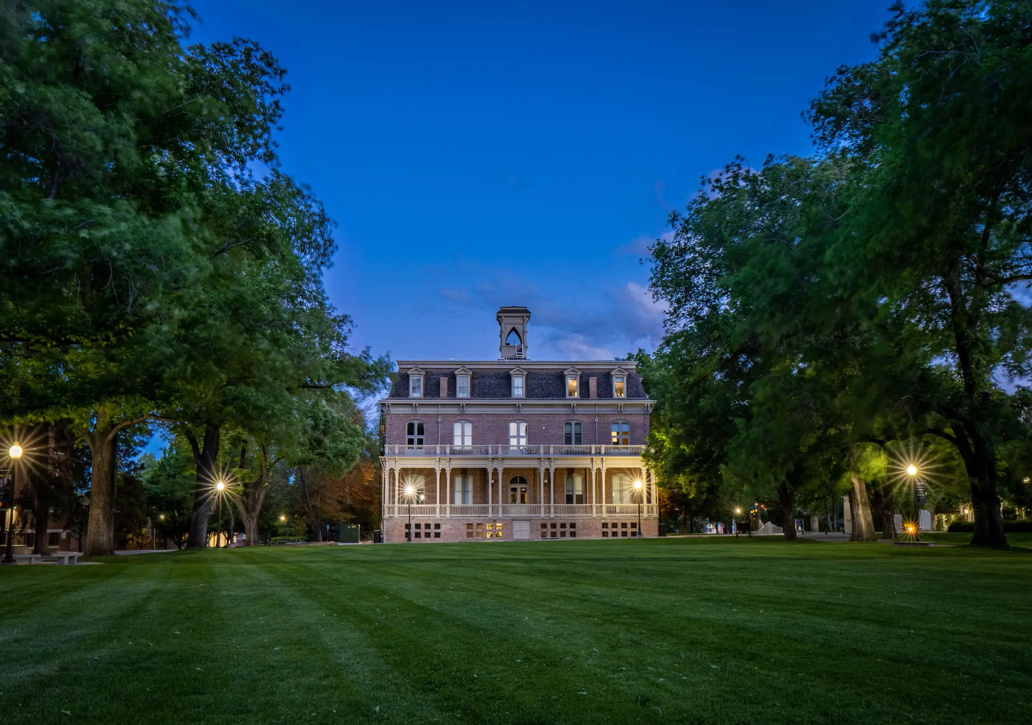 Historic brick building stands beyond a broad lawn framed by trees and glowing lights at dusk.