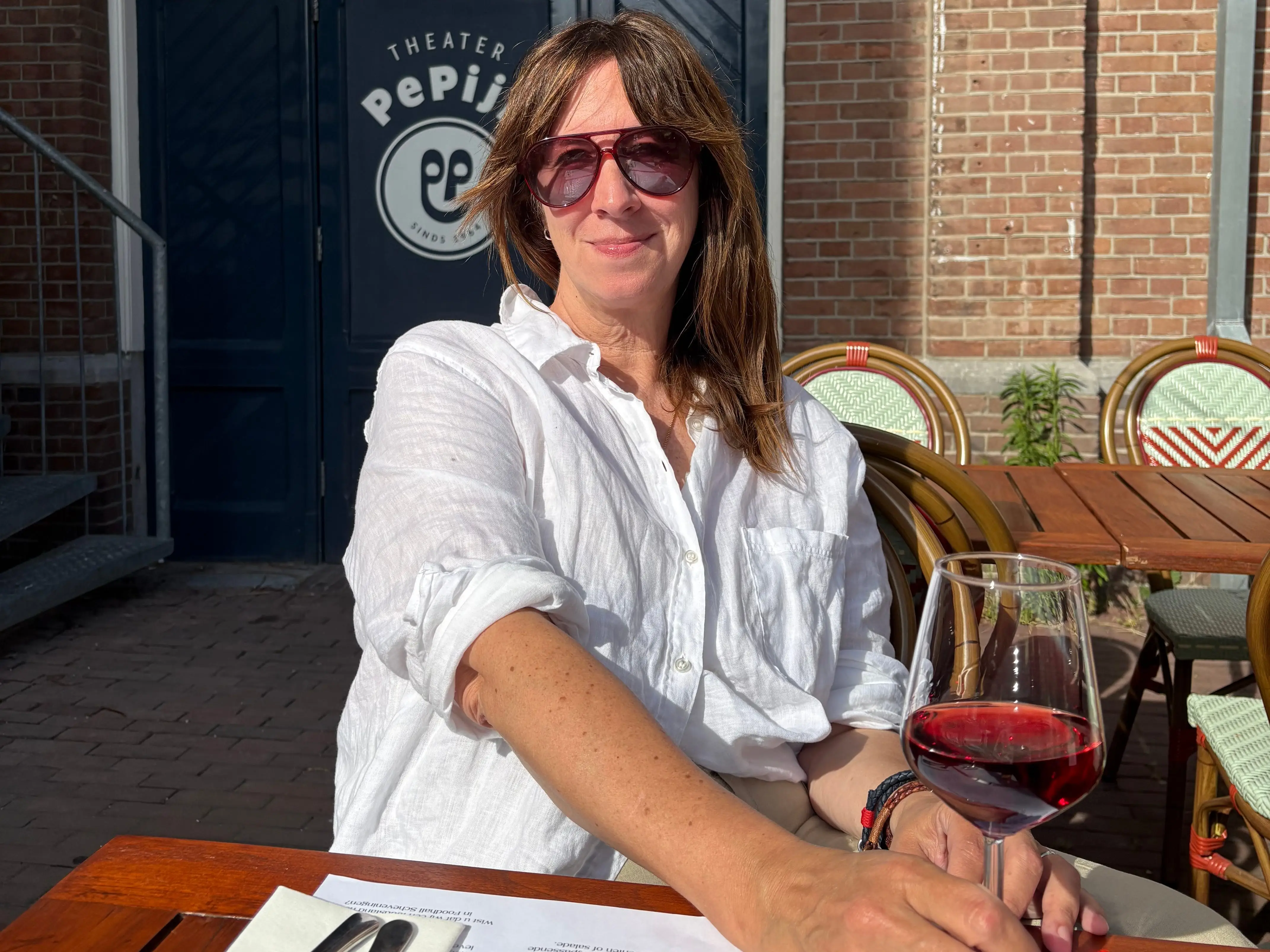 Person in sunglasses sits at an outdoor café table holding red wine outside Theater Pepijn.