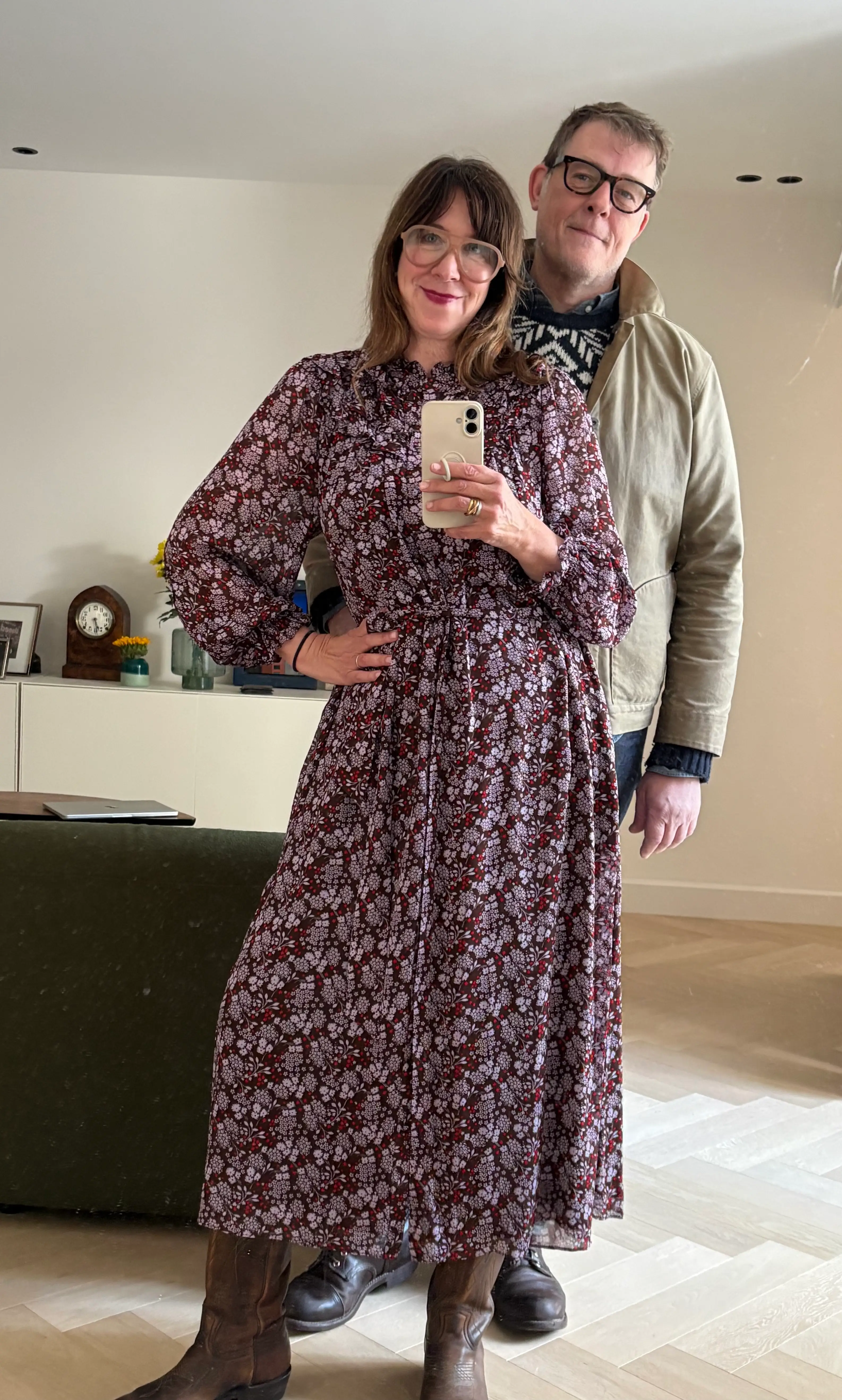 Two adults pose for a mirror selfie in a bright room with wood flooring and a sideboard behind them.