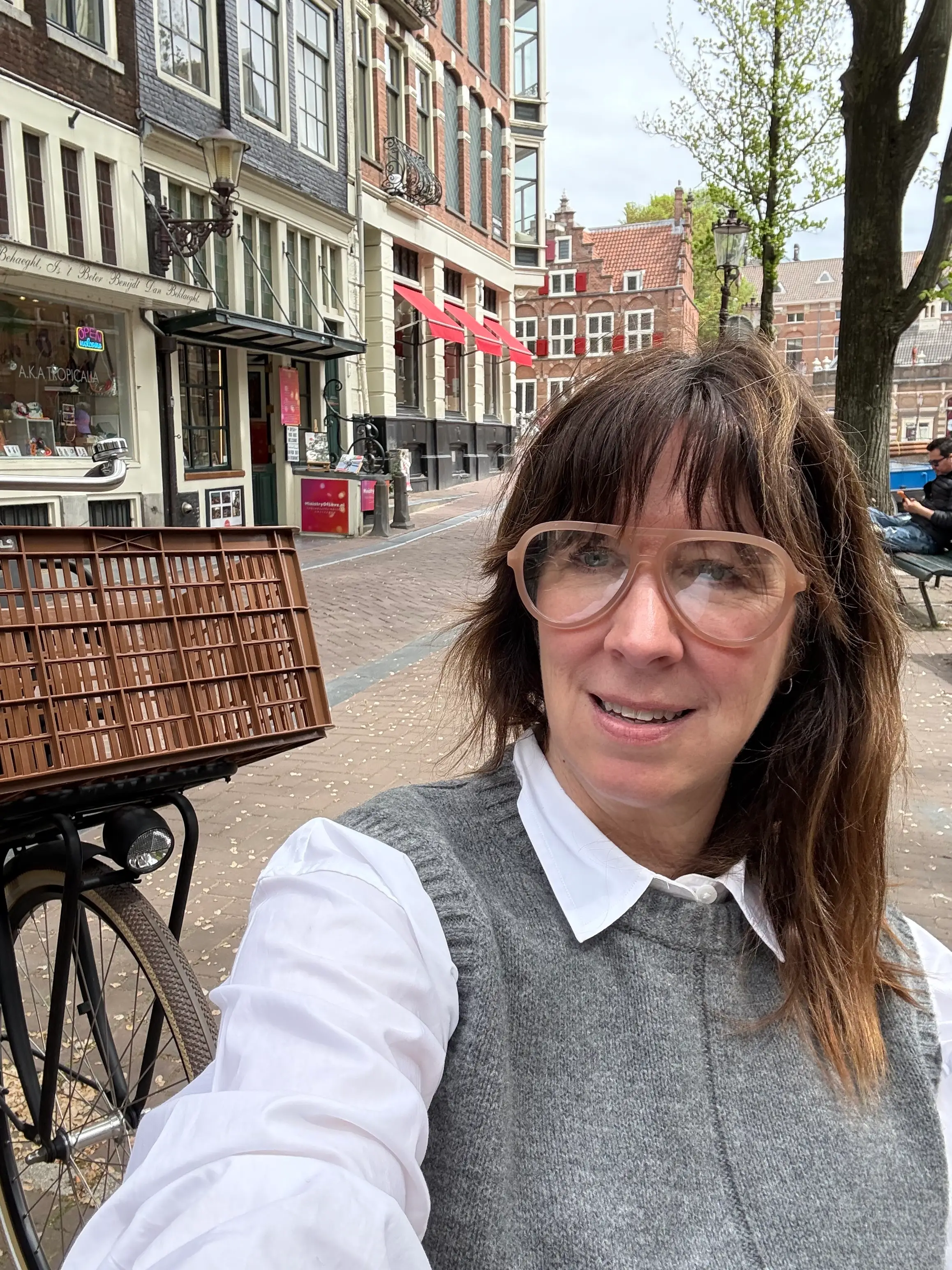 Person wearing large glasses takes a selfie on a cobblestone street beside a bicycle and historic storefronts.